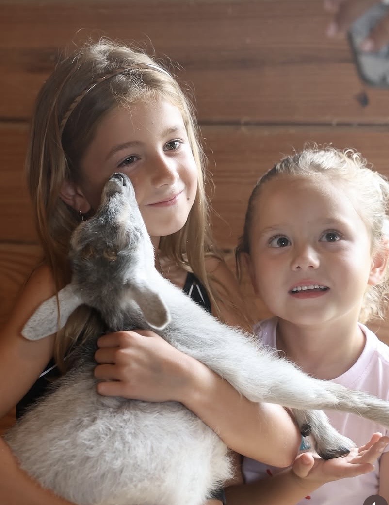 Two young girls holding a puppy indoors, with a wooden wall in the background.