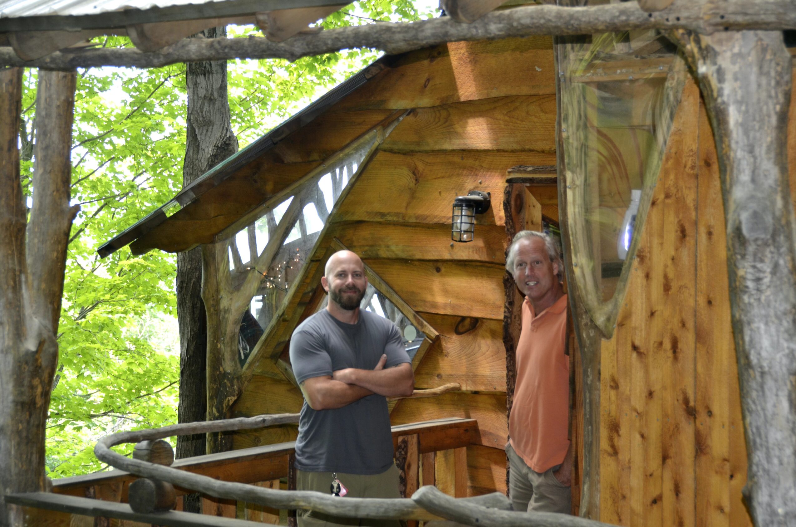 Two men stand outside a small wooden cabin in a forest, smiling at the camera.