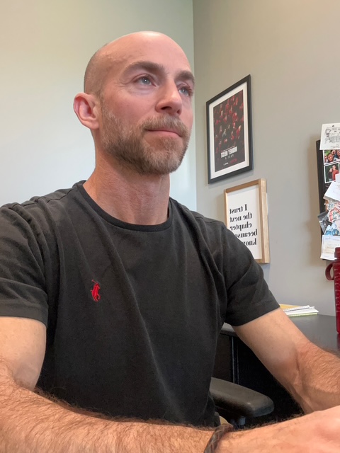 Man with a beard and bald head sitting indoors, wearing a black t-shirt, looking to the side.