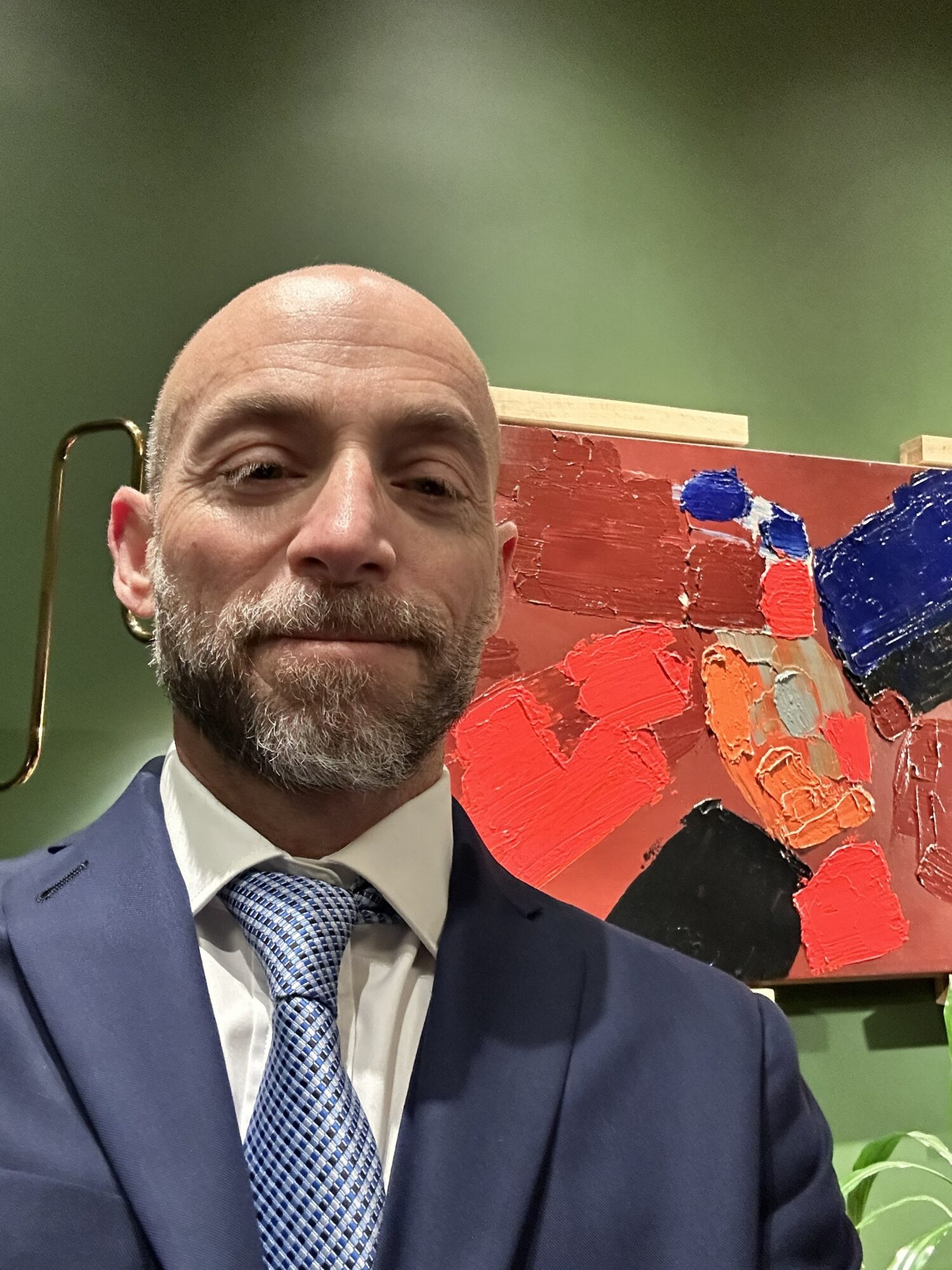 Man in suit and tie taking a selfie in front of an abstract painting with red, blue, and black colors.
