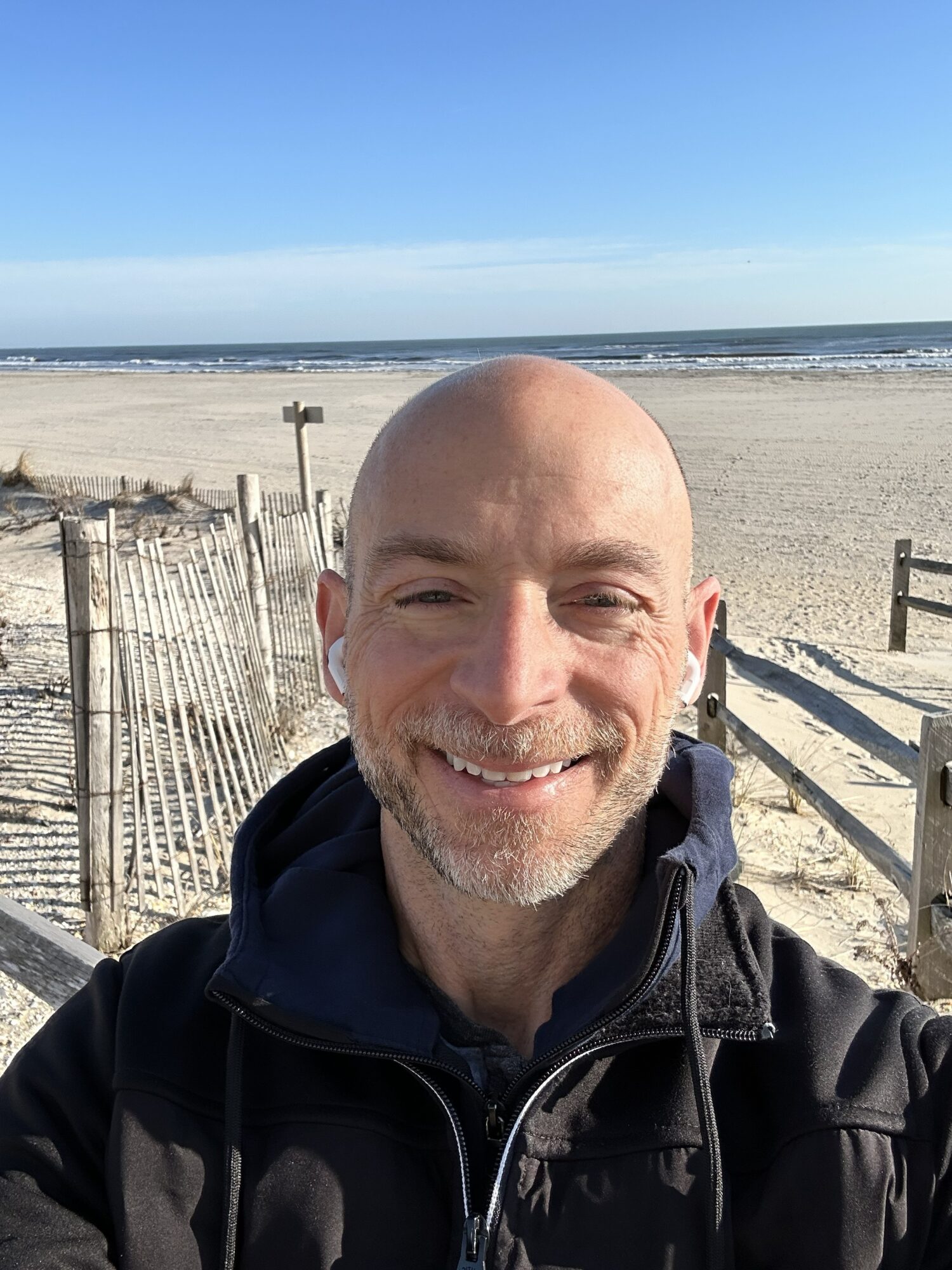 Man smiling at the beach with sand, fences, and ocean in the background.