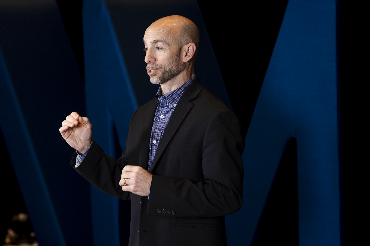 Man speaking on stage, gesturing with his right hand, wearing a dark blazer and blue shirt, with a dark background.