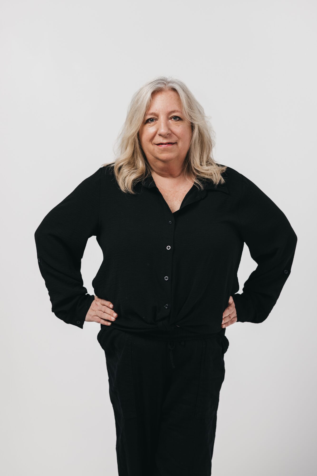 A woman with shoulder-length gray hair wearing a black outfit, standing with hands on hips, smiling.