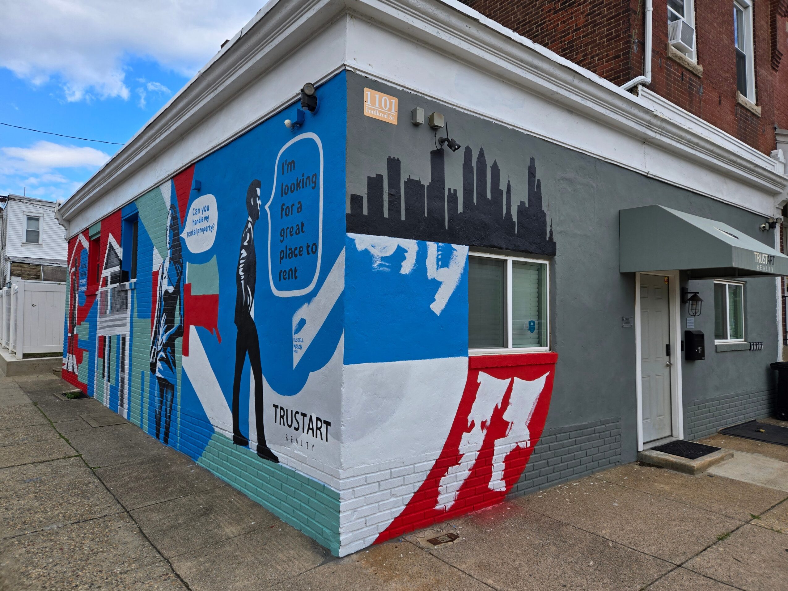 Corner of a building with colorful mural, city skyline silhouette, and a person holding a phone, with a sidewalk in front.