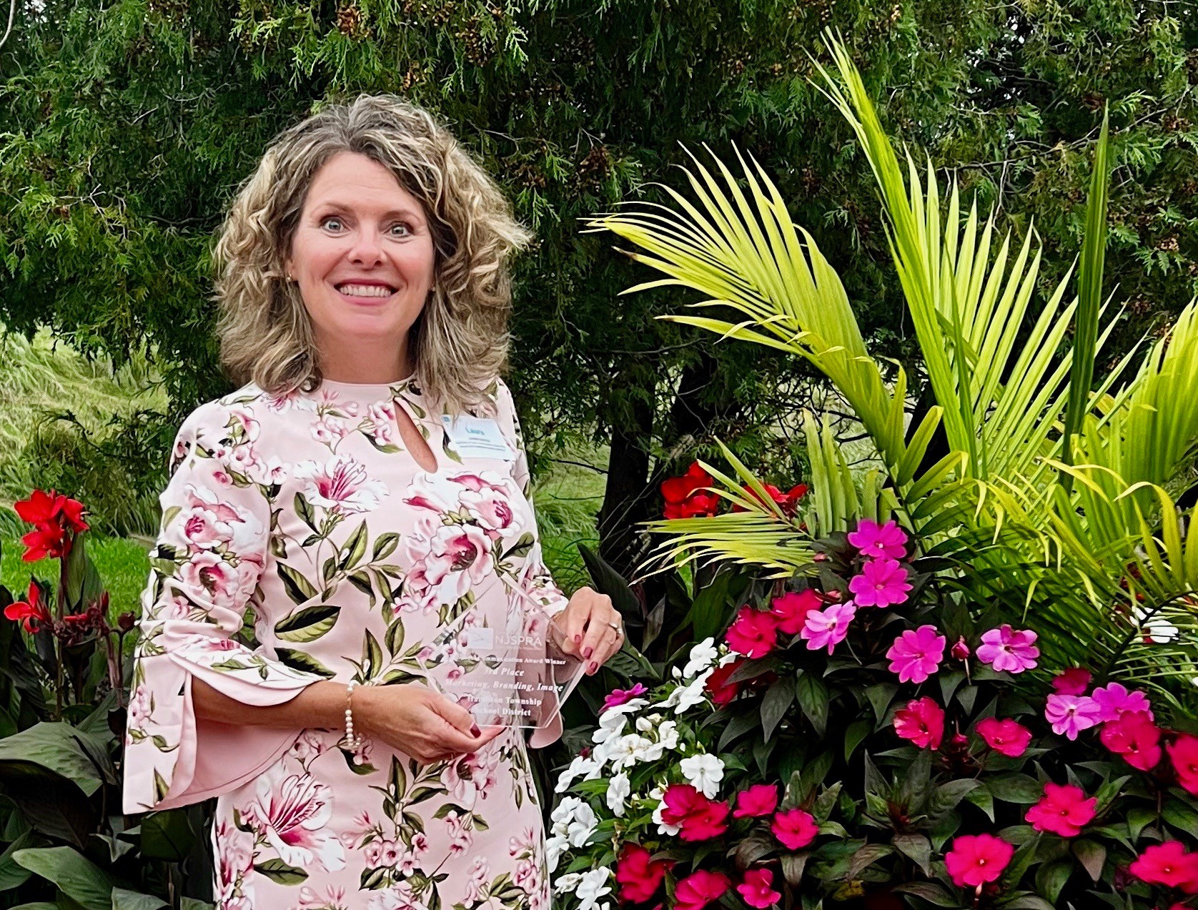 Woman with curly hair in floral dress standing outdoors with green plants and colorful flowers.