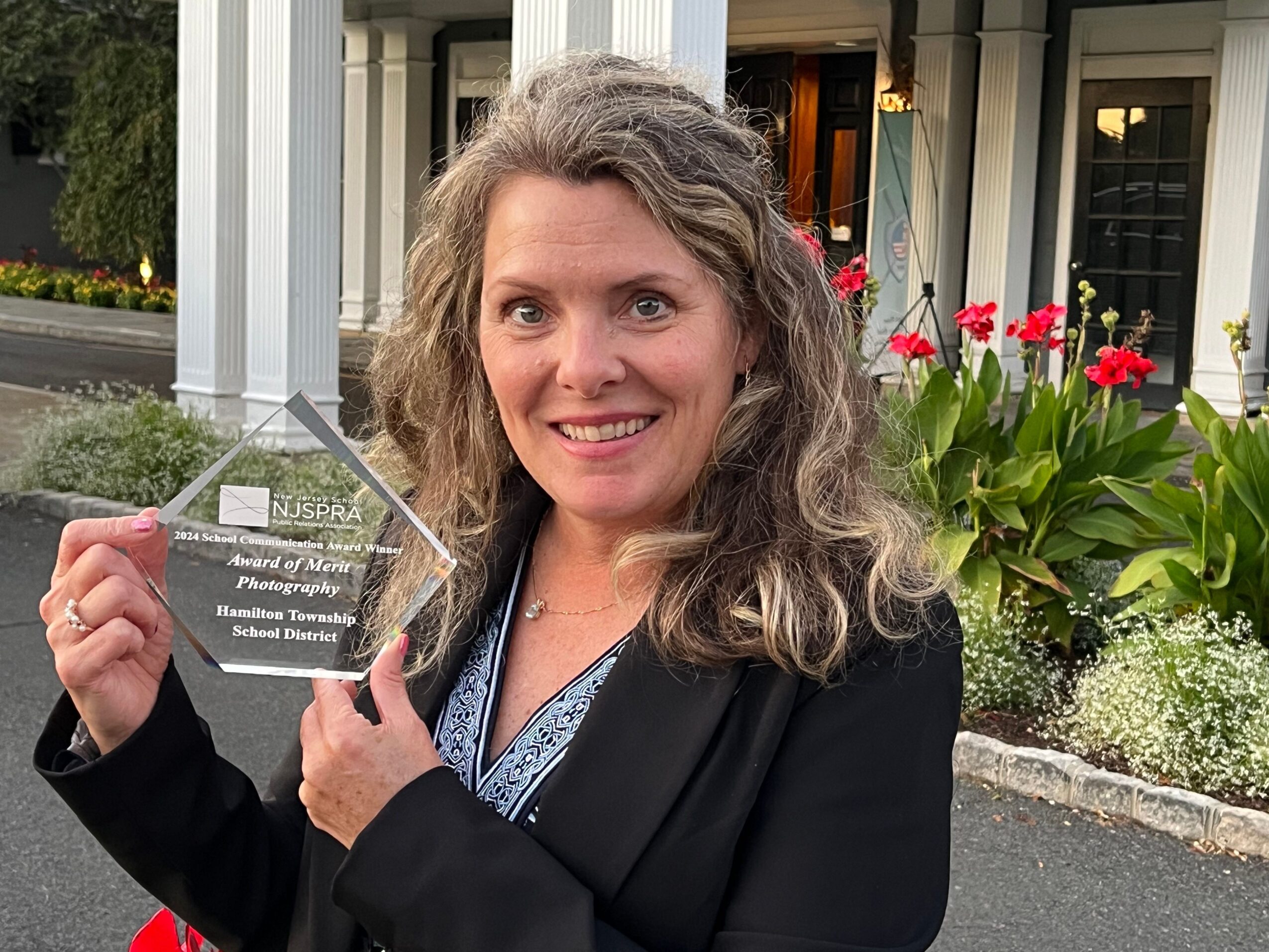 Woman with curly hair smiling, holding a glass award, standing outdoors near a plant with red flowers.