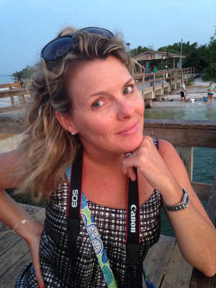 Woman with sunglasses on her head, smiling, sitting outdoors near water with a wooden dock and buildings in background.