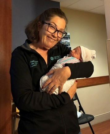 Woman holding a newborn baby, smiling, in an indoor setting, wearing glasses and a dark jacket.