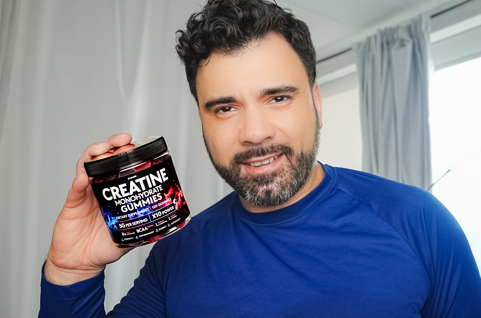 Man with dark curly hair and beard holding a container of creatine gummies, smiling, indoors with curtains and window behind him.