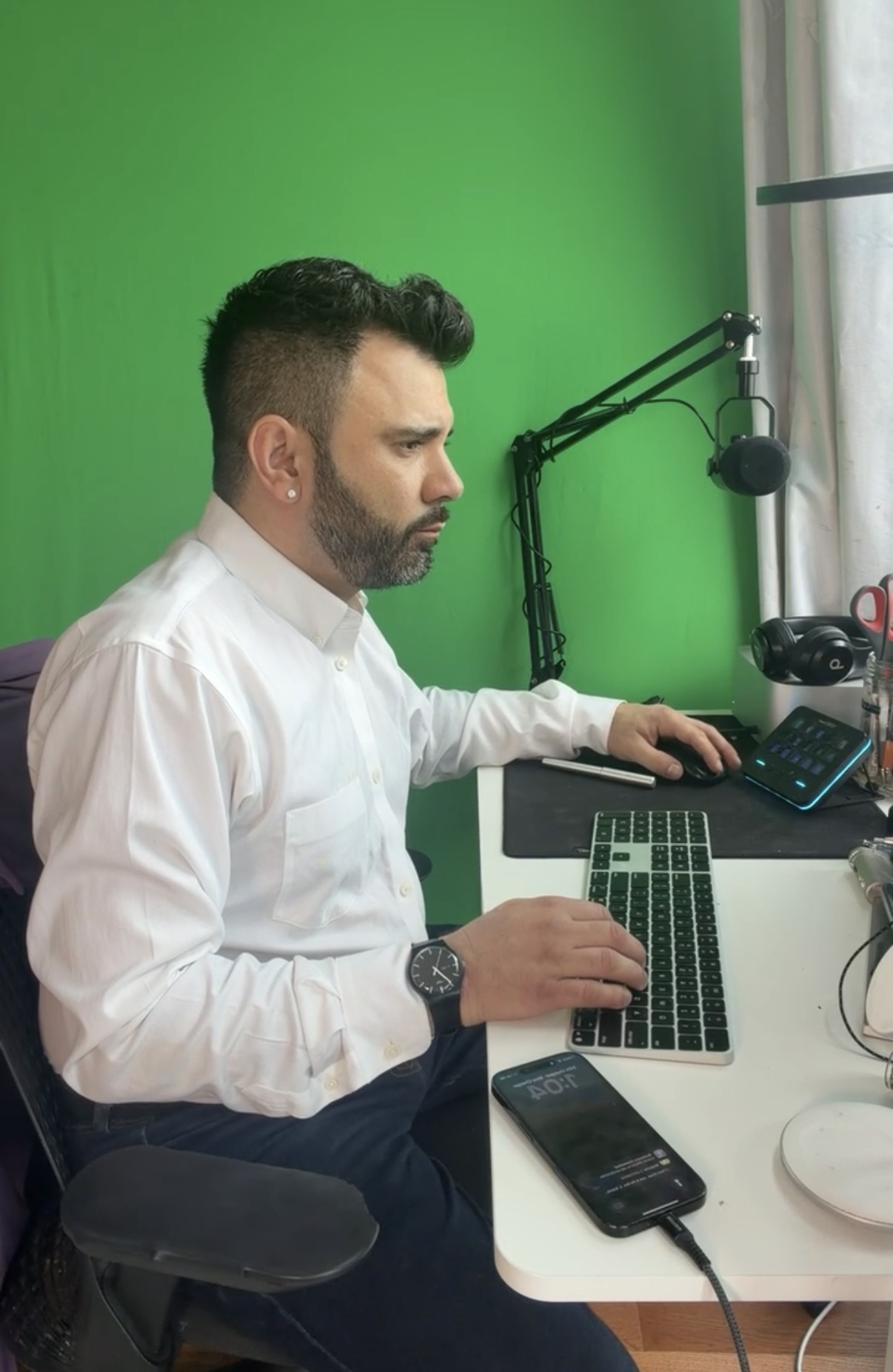 Man with dark hair and beard in white shirt sitting at desk with microphone, keyboard, and phone, against green wall.