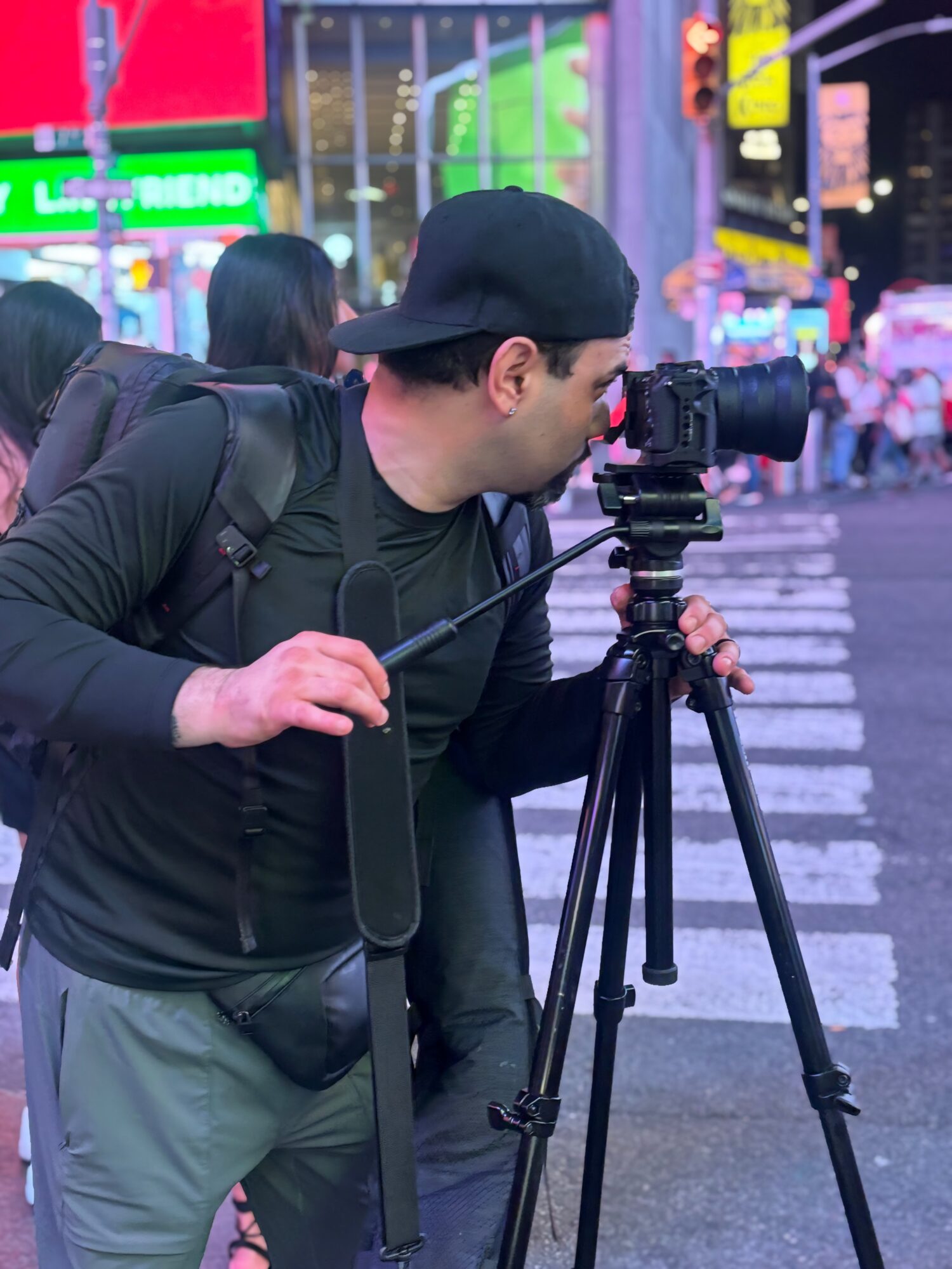 Person with a backpack operating a camera on a tripod in a city at night with bright neon lights