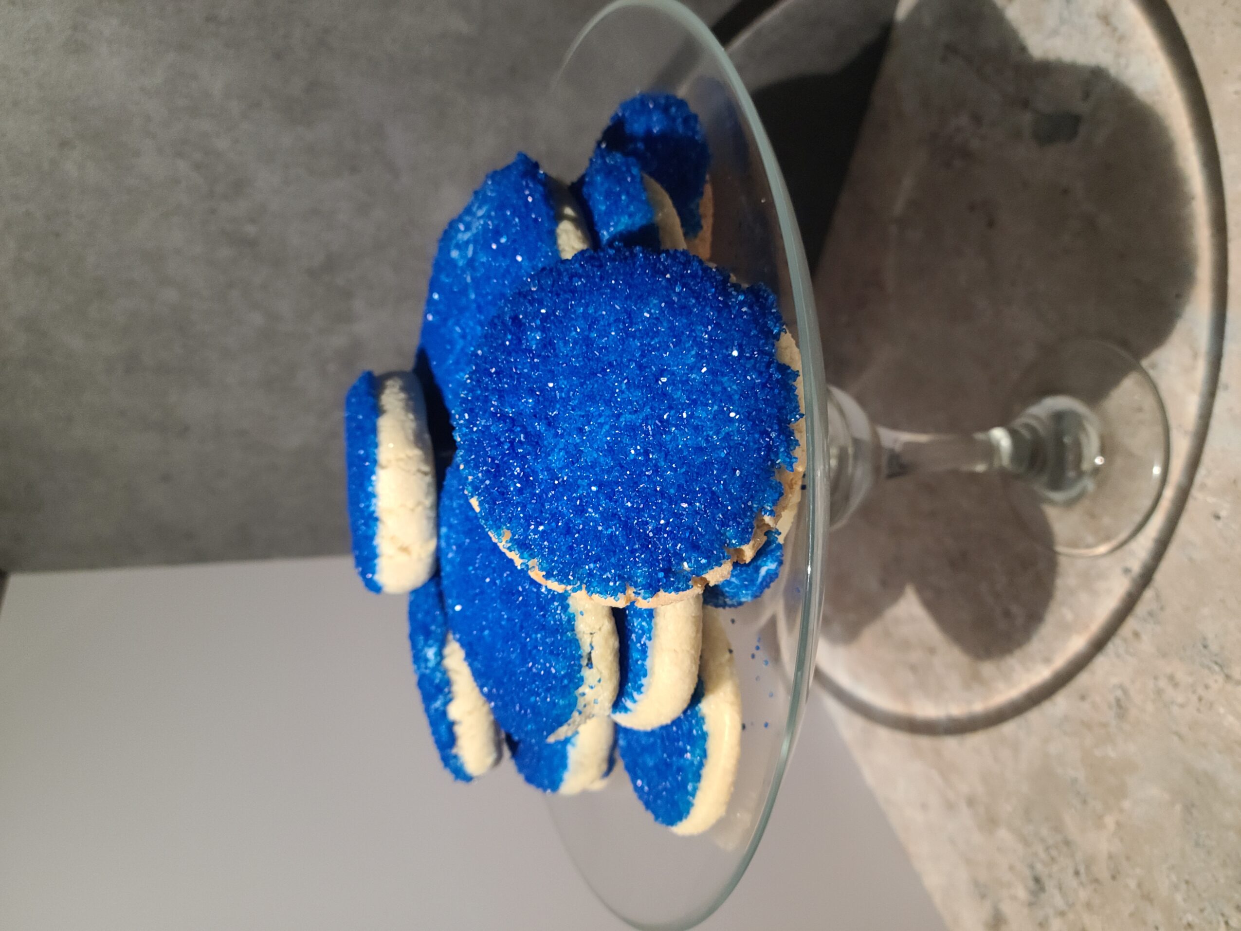 Blue cookies with white filling stacked on a glass cake stand, set on a gray surface.