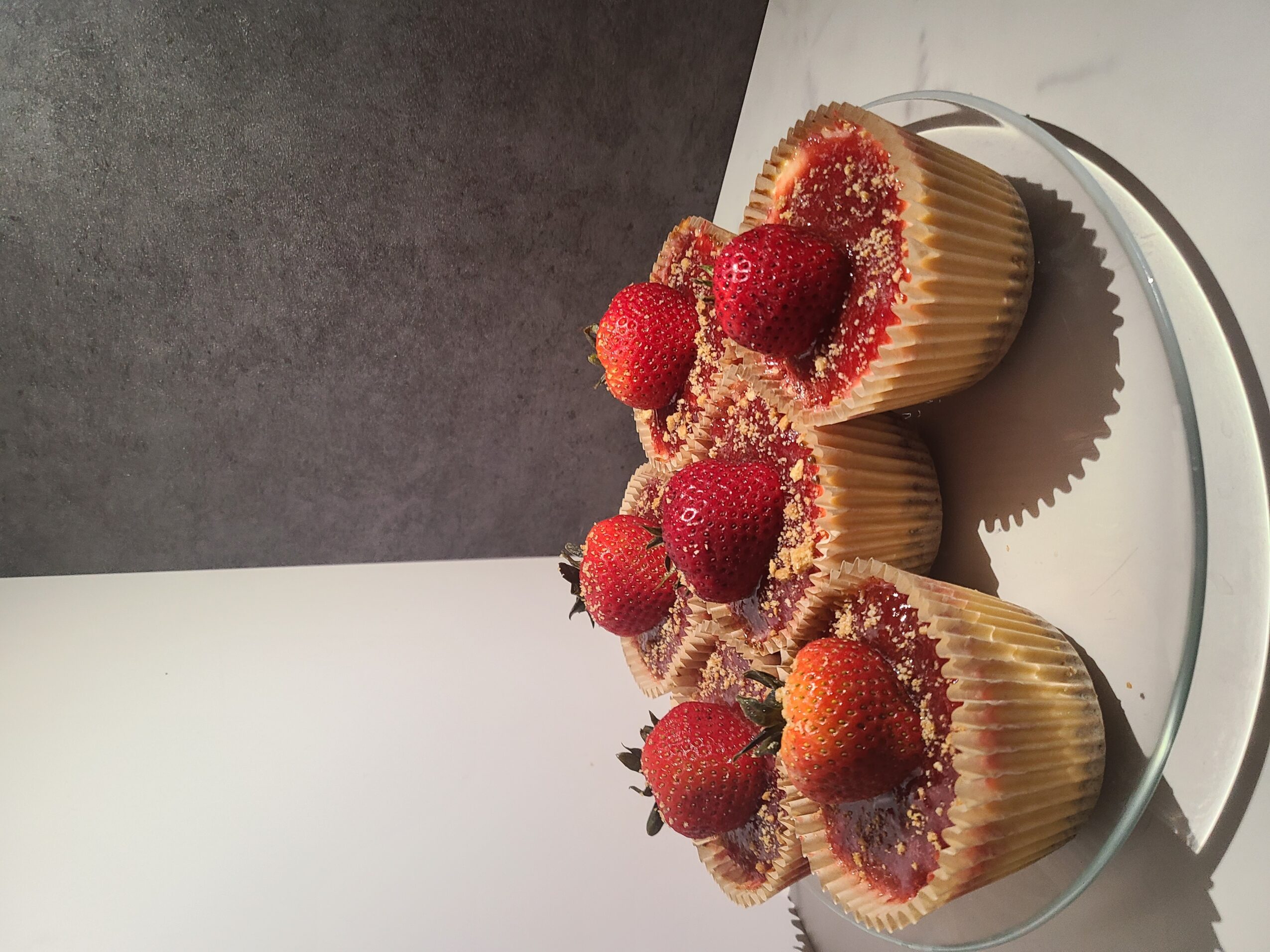 Three cupcakes topped with strawberries and sprinkles on a glass plate, against a dark and light background.