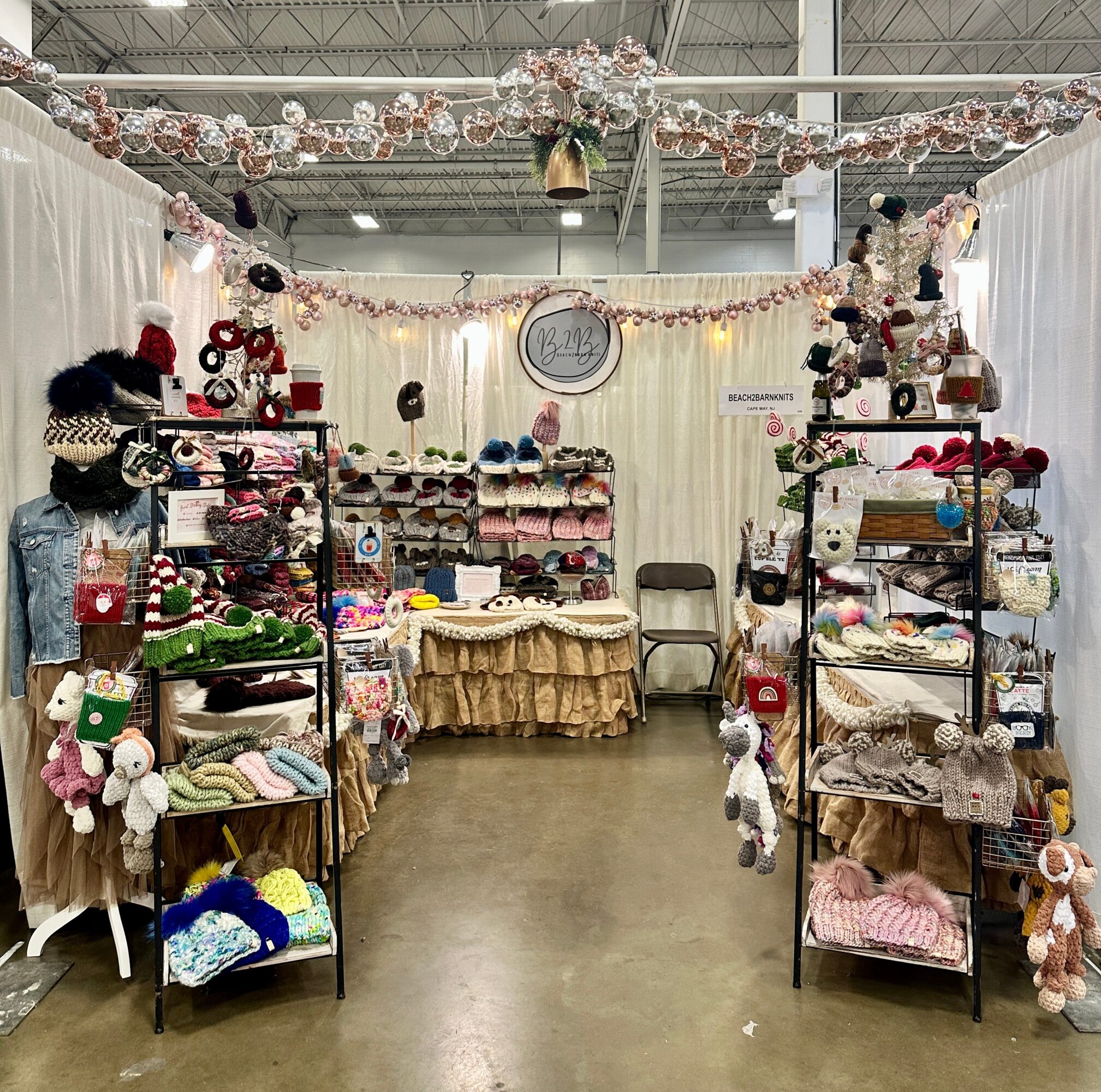 Indoor craft booth with shelves of handmade items, decorated with garlands and a central table, against white curtains and a ceiling with string lights.
