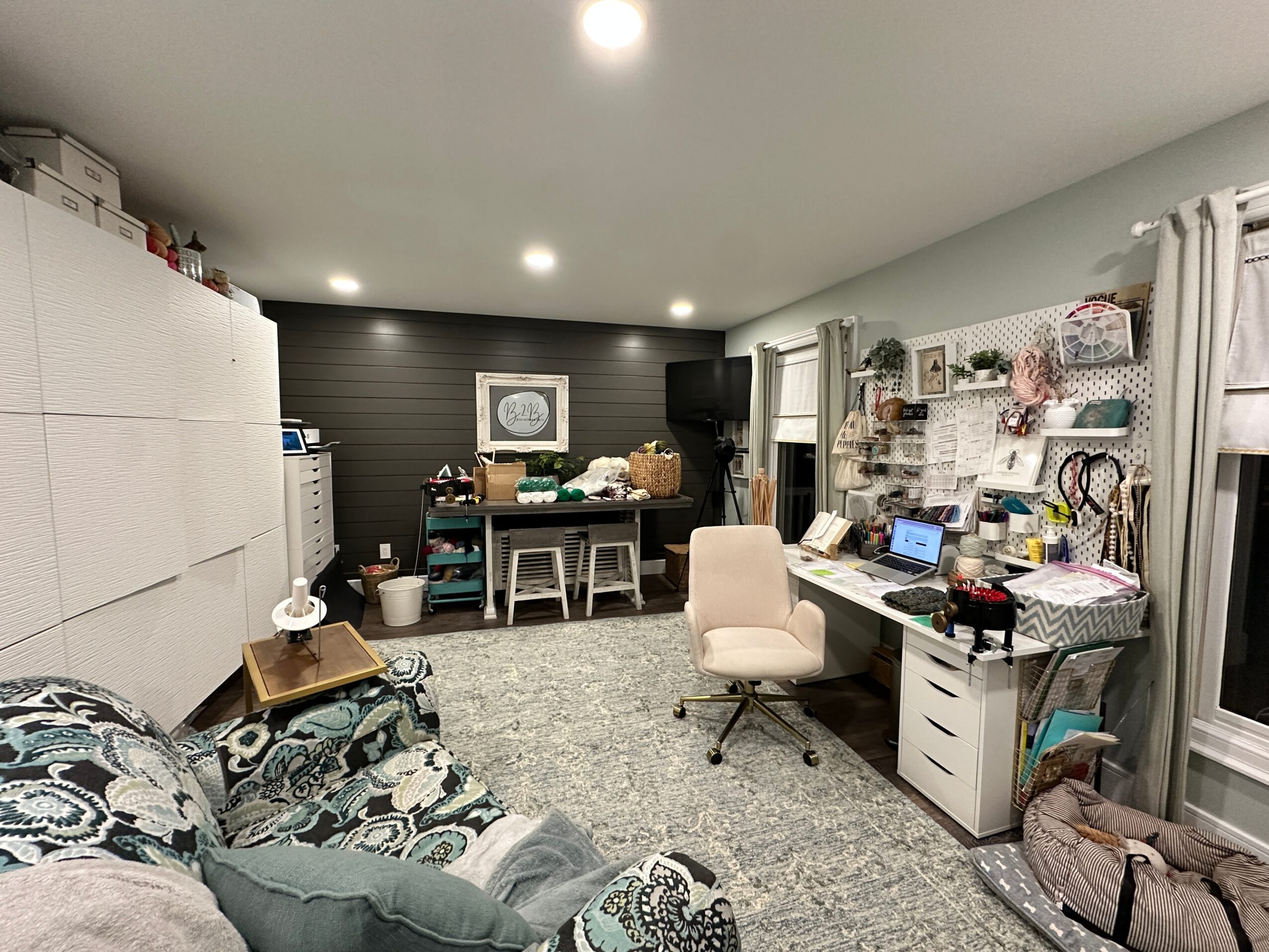 Home office with desk, chair, shelves, and a sofa in a well-lit room with gray walls and a window.