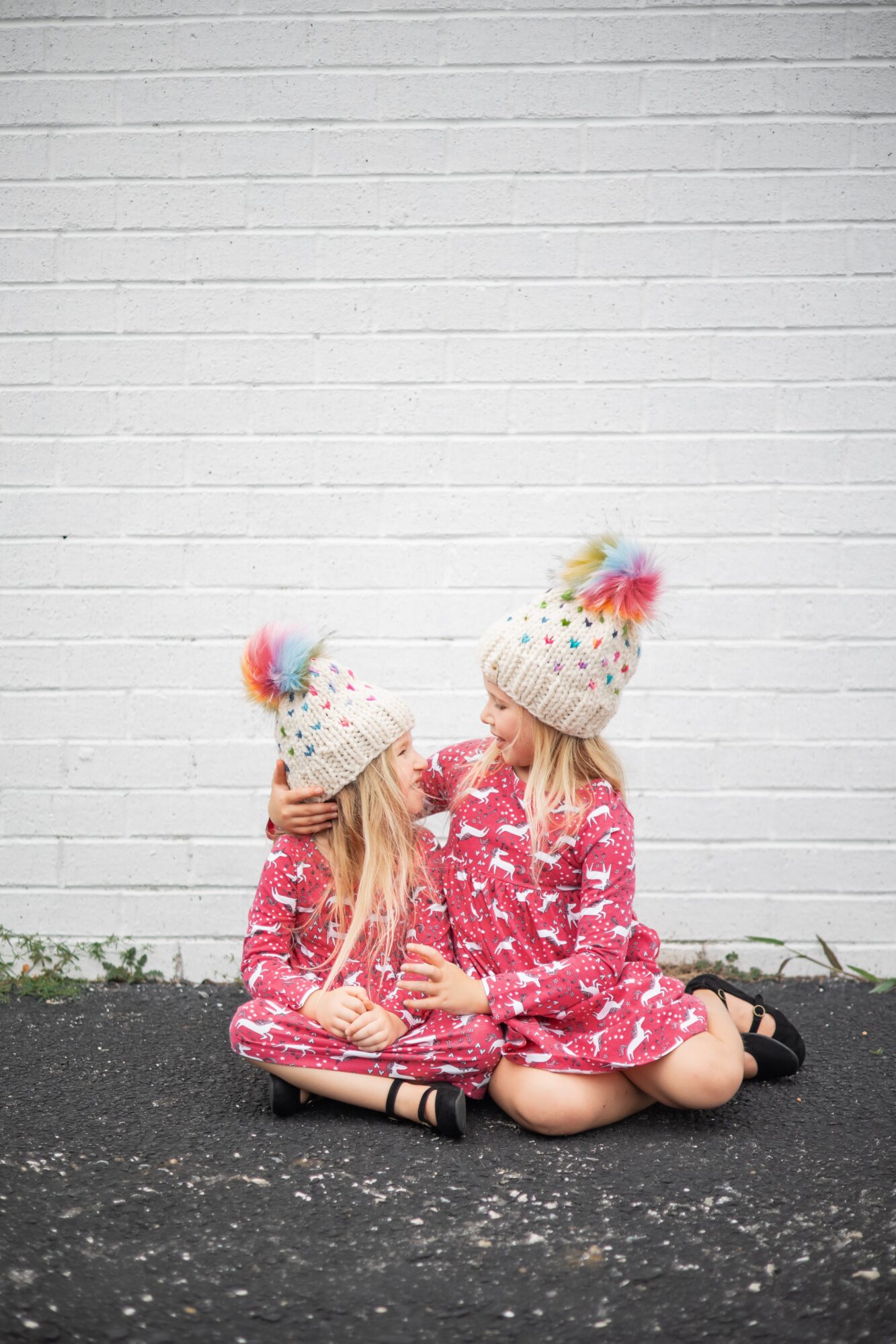 Two children sitting on the ground in front of a white brick wall, wearing matching pink dresses and colorful pom-pom hats, holding hands.