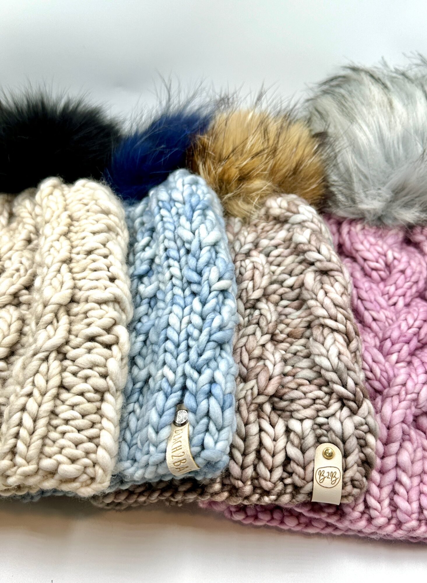 Four knitted winter hats with pom-poms, in beige, blue, gray, and pink, arranged side by side on a white surface.