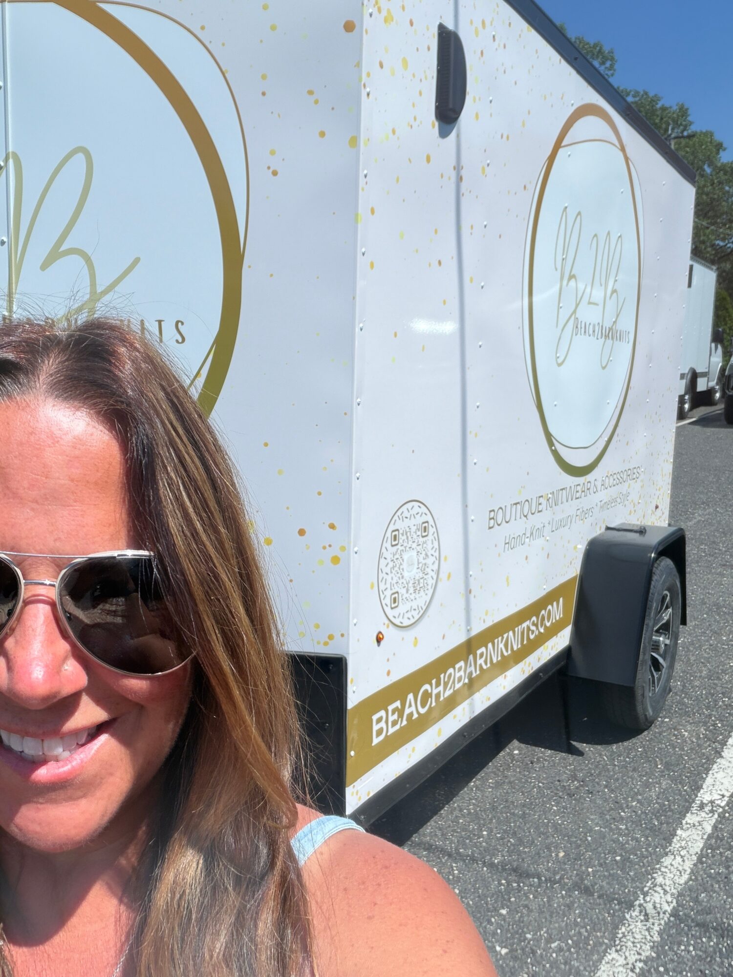 Part of a woman with sunglasses and long hair, smiling, standing next to a white vehicle with logo and text, outdoors.