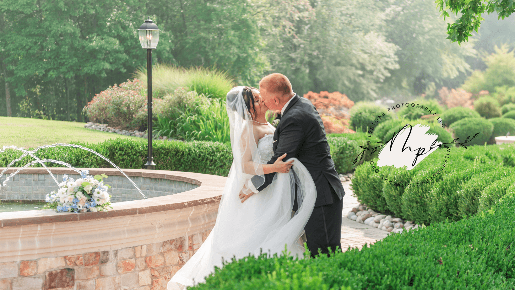 A bride and groom embrace in a garden with greenery and a fountain, during daytime. The bride wears a white dress and veil.