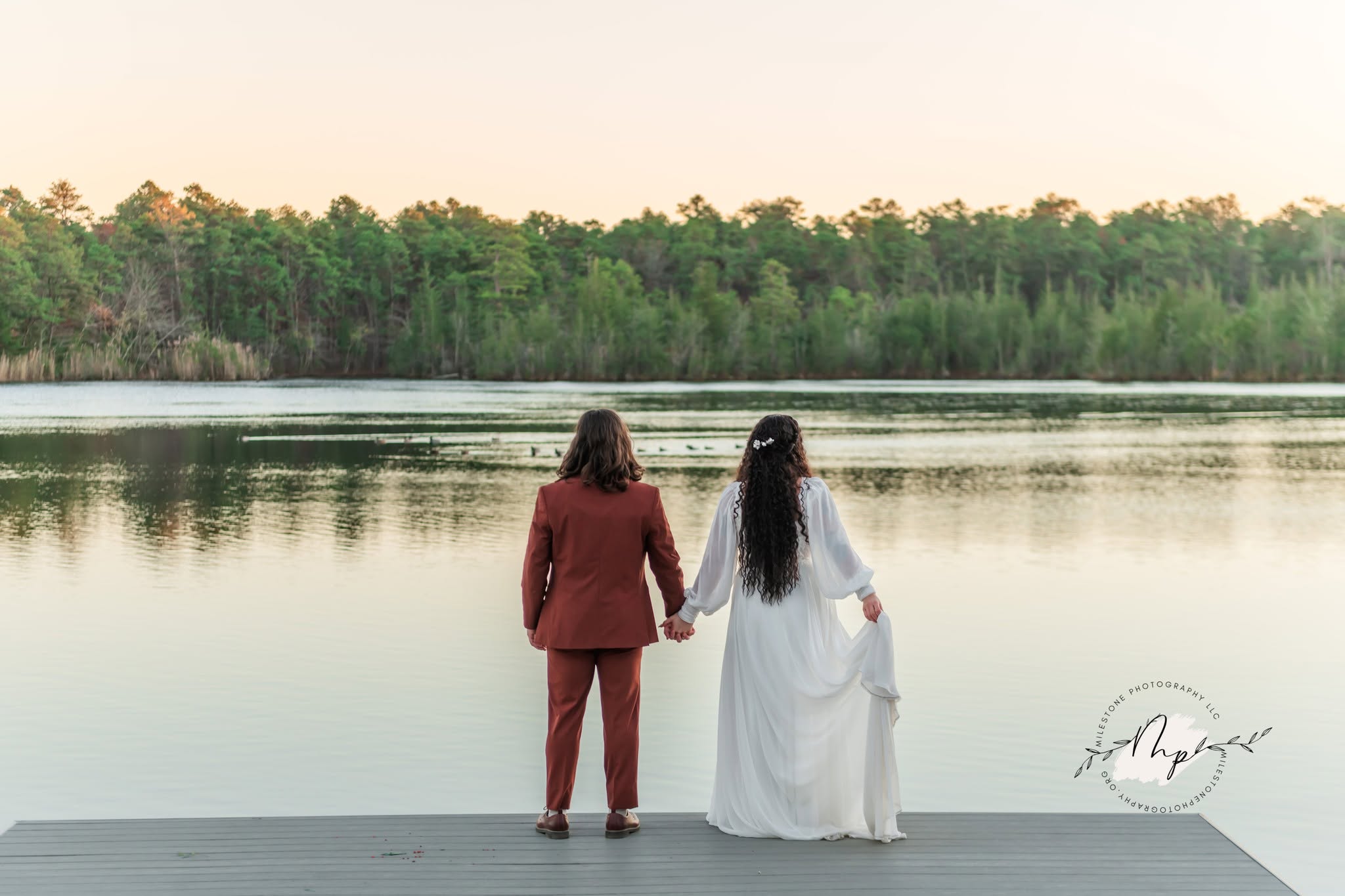 Two women hold hands by a lake with trees in the background, one in a red suit and the other in a white dress.