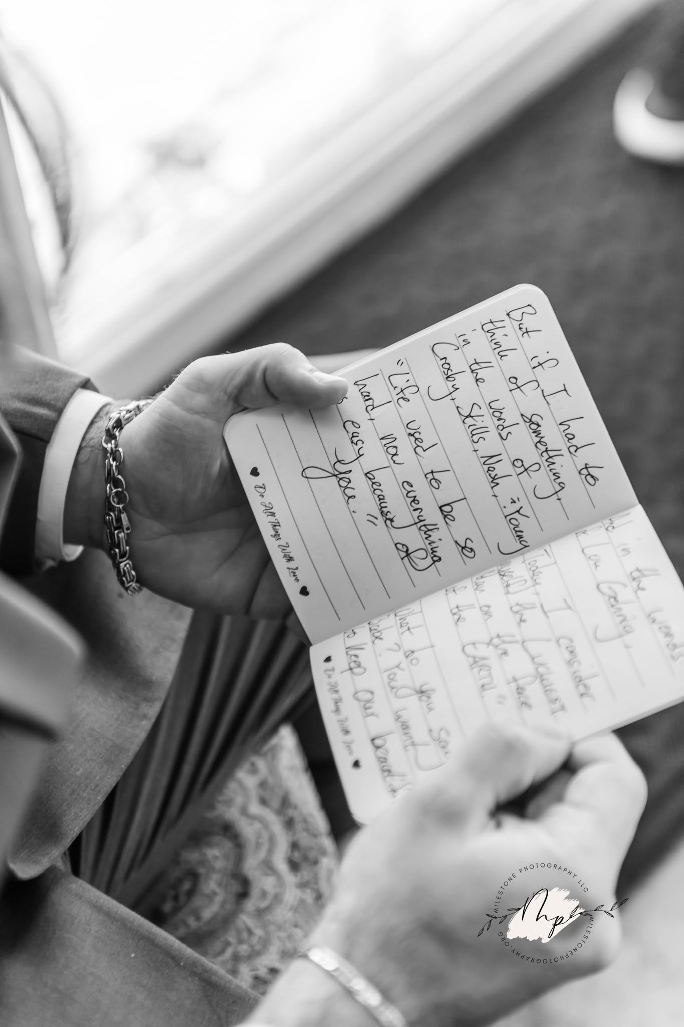 Person holding an open notebook with handwritten notes, wearing bracelets, near a window with a blurred background.