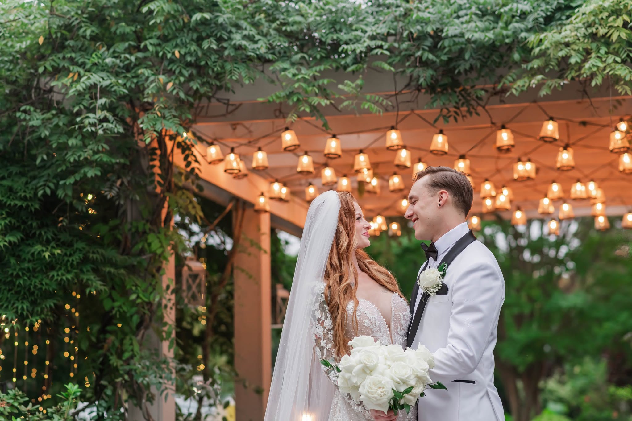Bride and groom face each other under decorated canopy, surrounded by greenery, holding flowers, during wedding ceremony.