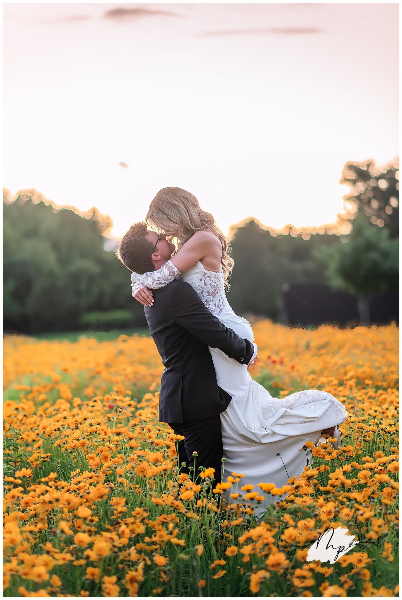 A couple embraces in a field of yellow flowers at sunset, with trees in the background.