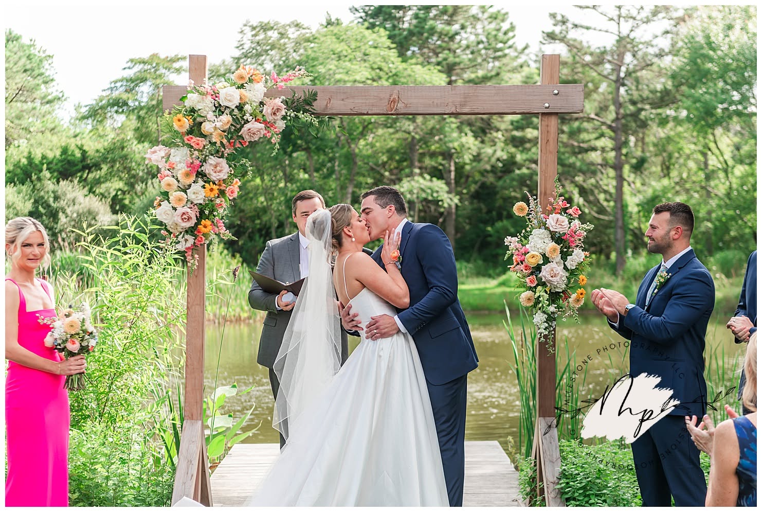 Couple kissing during wedding ceremony outdoors with floral arch, surrounded by guests, greenery, and trees.
