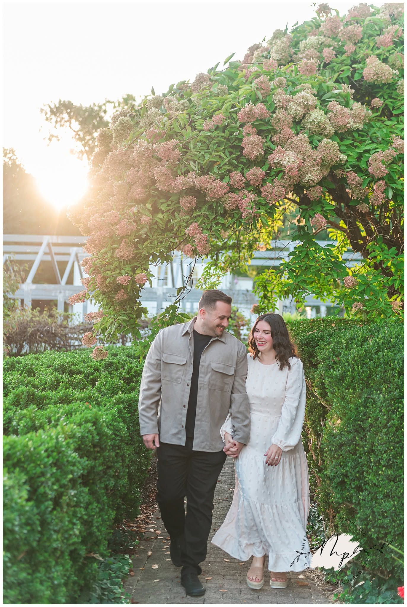 A man and woman walk hand in hand outdoors, surrounded by greenery and a flowering tree, with sunlight in the background.