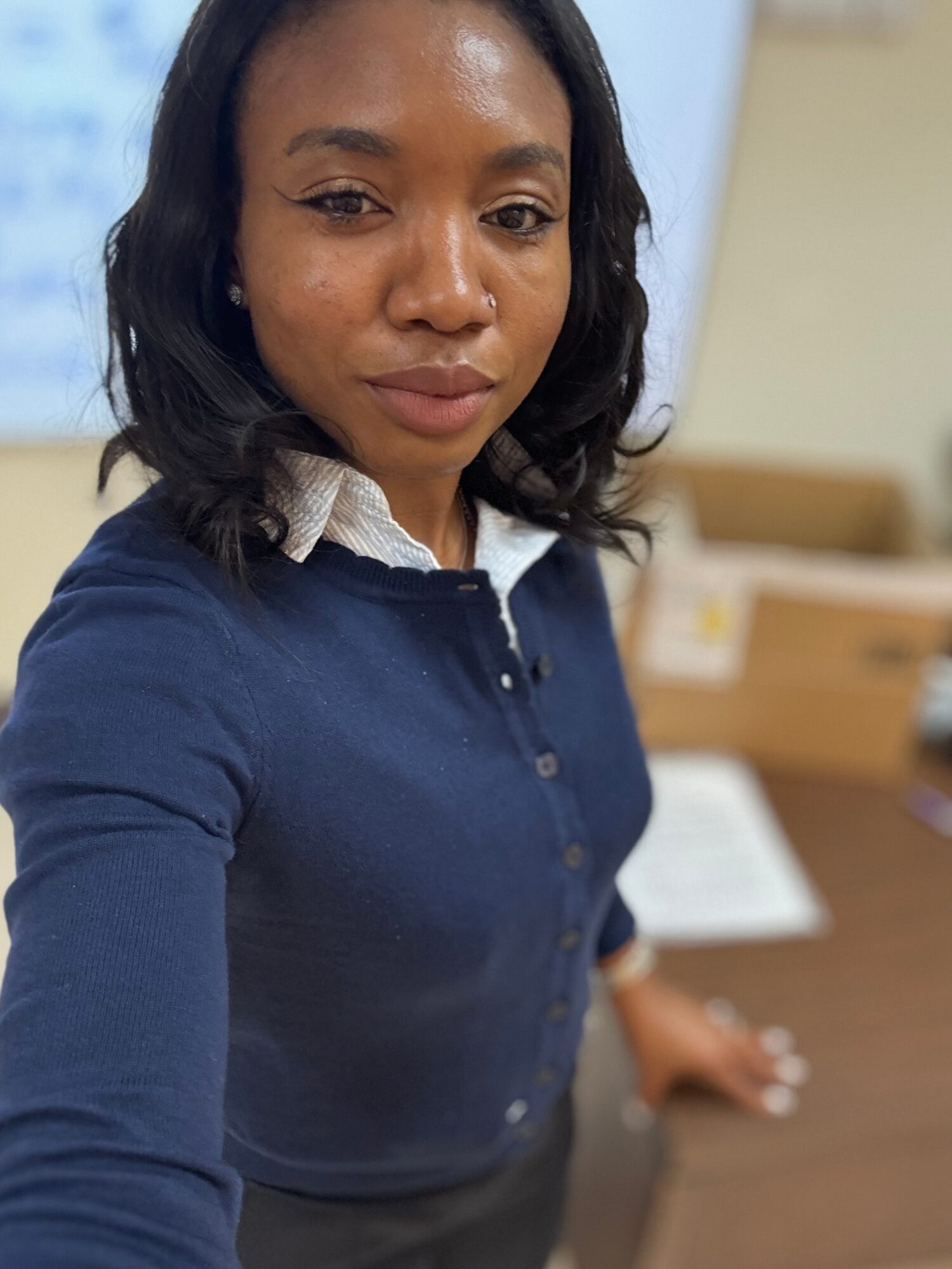 Woman with dark hair wearing a navy blue cardigan and white collared shirt, standing indoors with blurred background.
