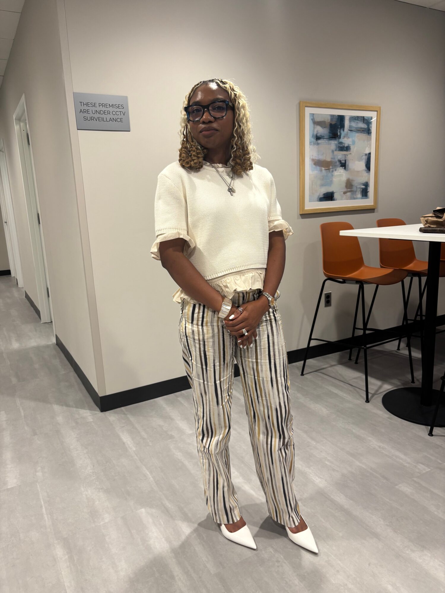 Woman with curly hair and glasses stands in a room with artwork on the wall, wearing a white top and striped pants.