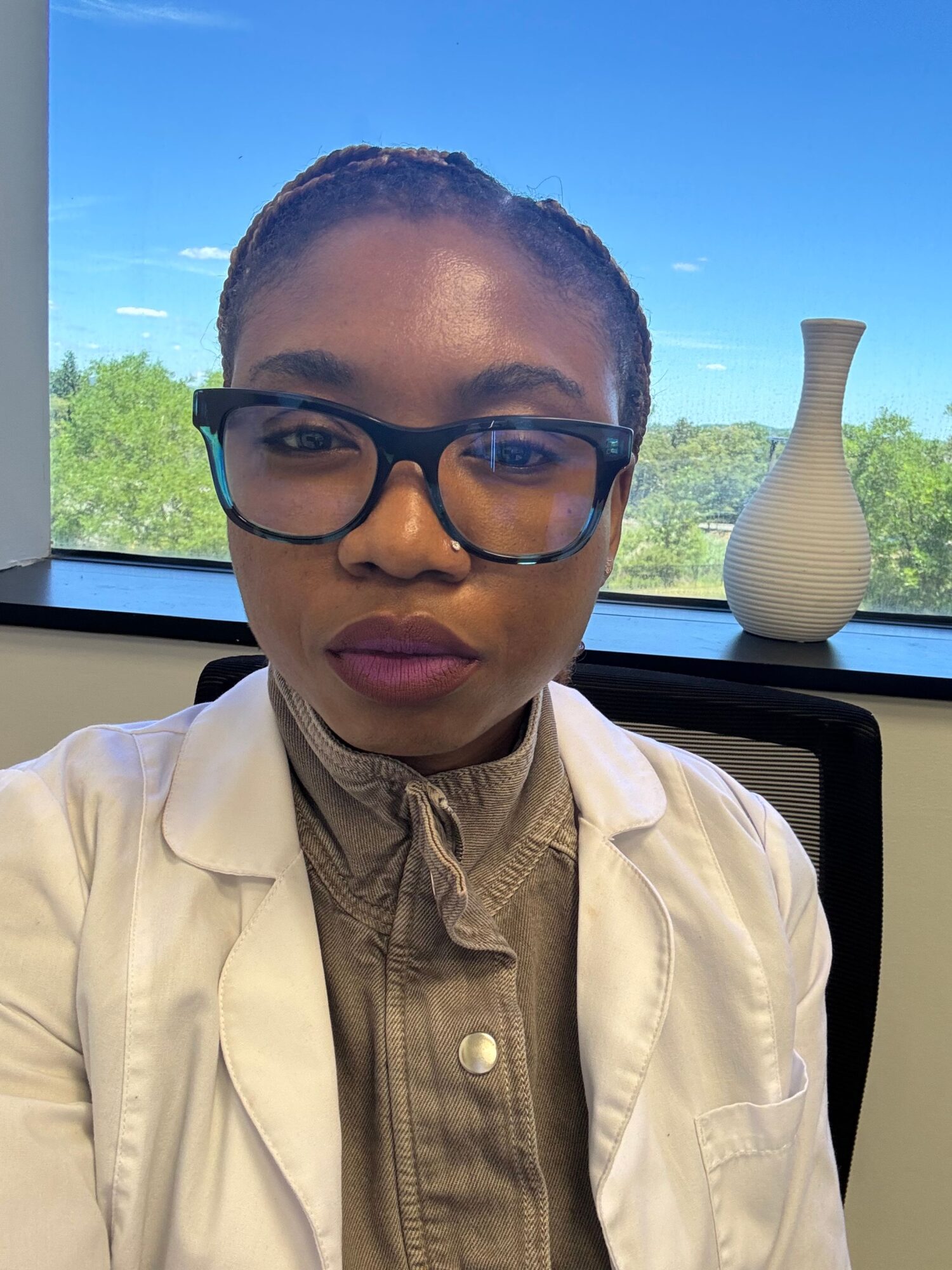Person with glasses and braided hair, wearing a white jacket and beige shirt, sitting near a window with a vase outside.