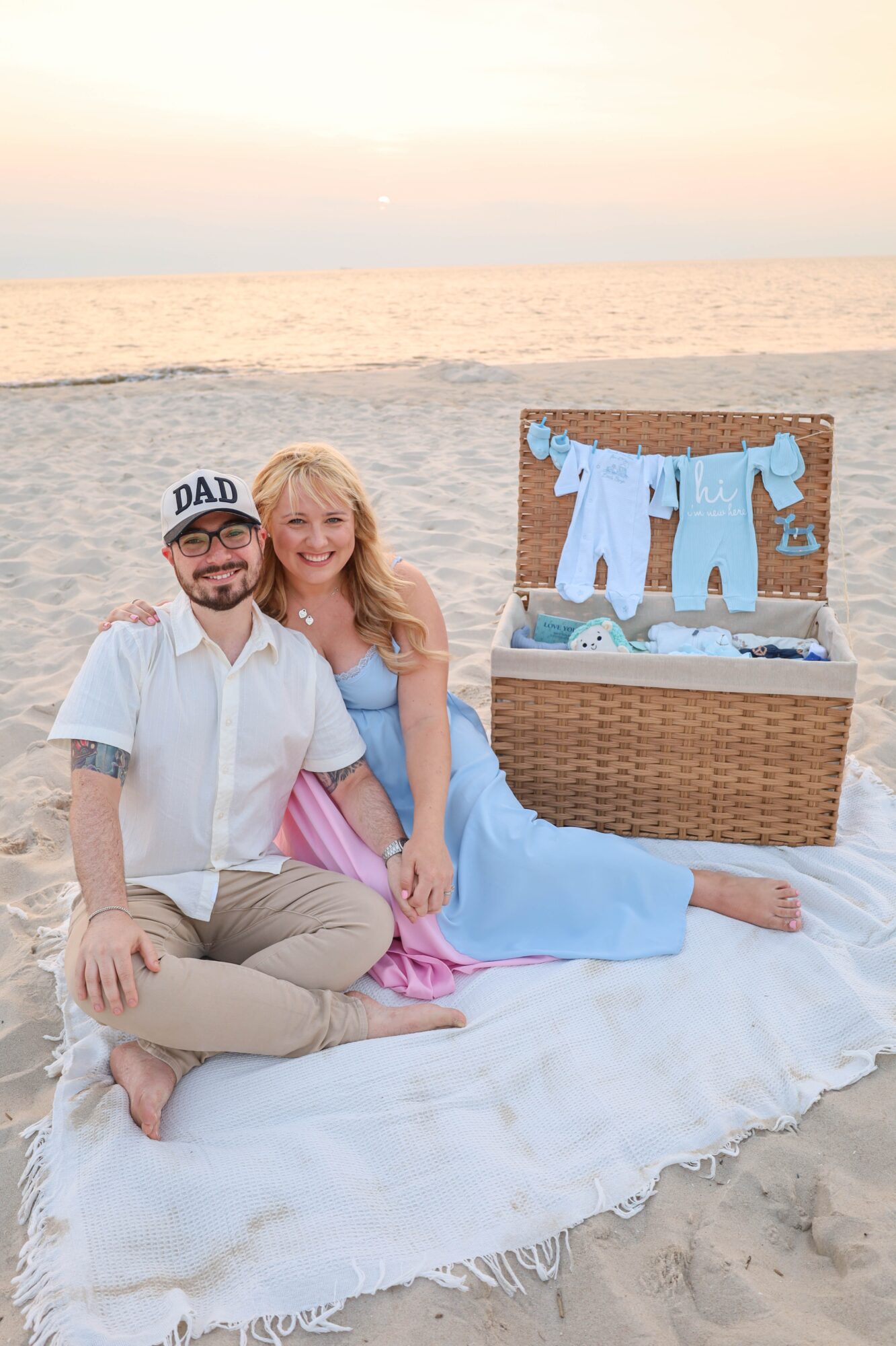 A couple sitting on a blanket at the beach during sunset, with a picnic basket and clothes hanging inside it.