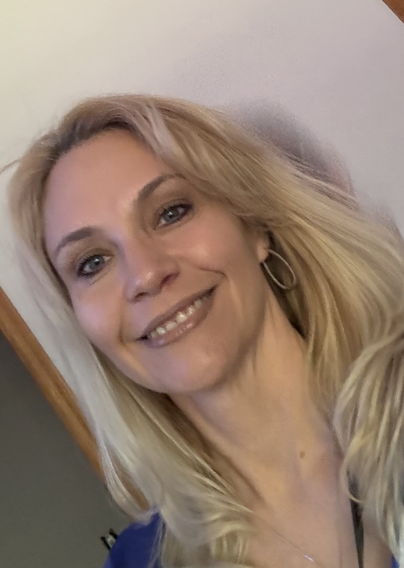 Smiling woman with blonde hair and hoop earrings, facing slightly to the right, against a plain background.