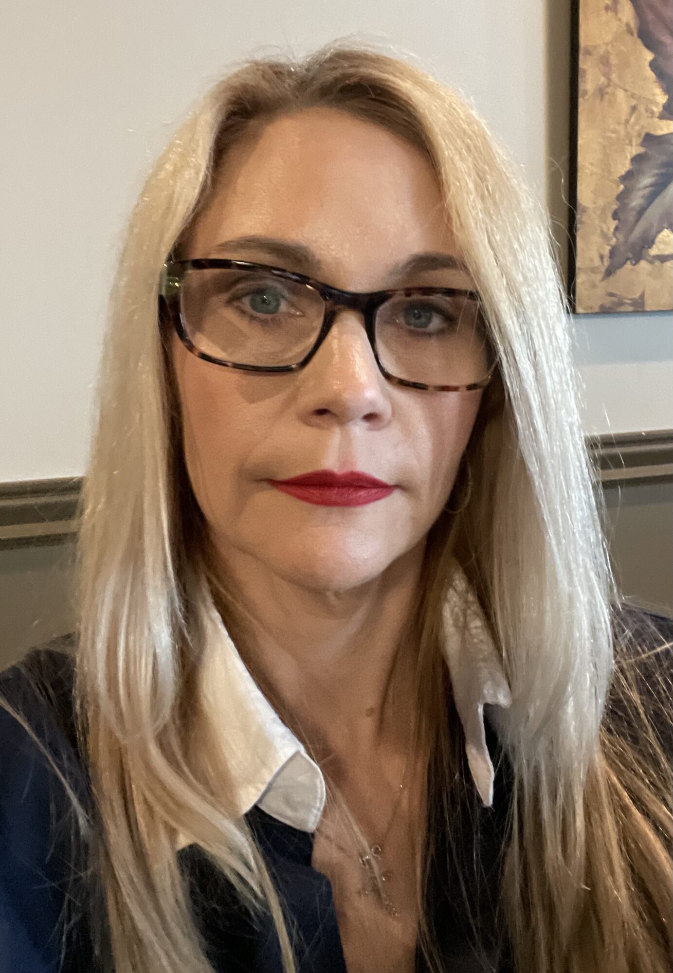 Woman with blonde hair, glasses, and red lipstick, wearing a white shirt and dark blazer, indoors.
