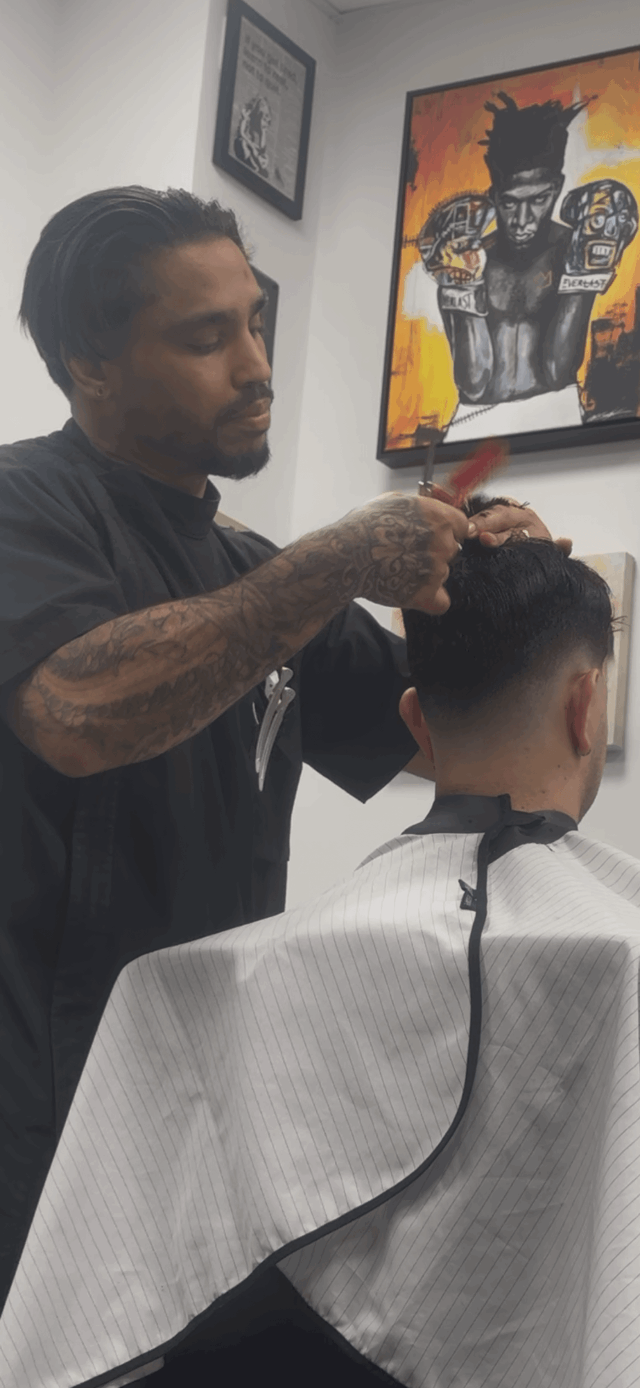 Barber cutting a person's hair in a salon with colorful artwork on the wall behind them.