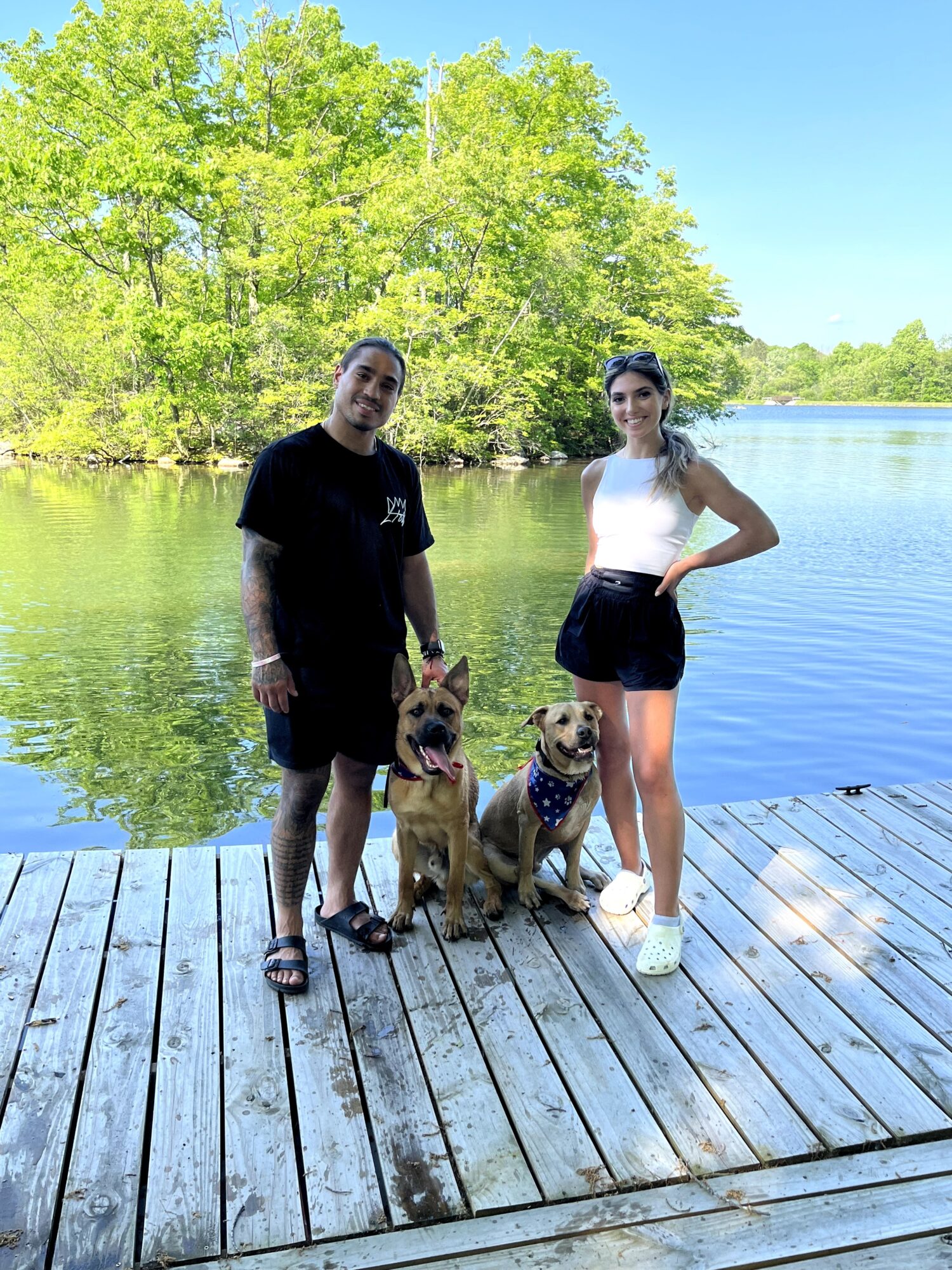 Two people and two dogs on a wooden dock by a lake with green trees and blue sky in the background.