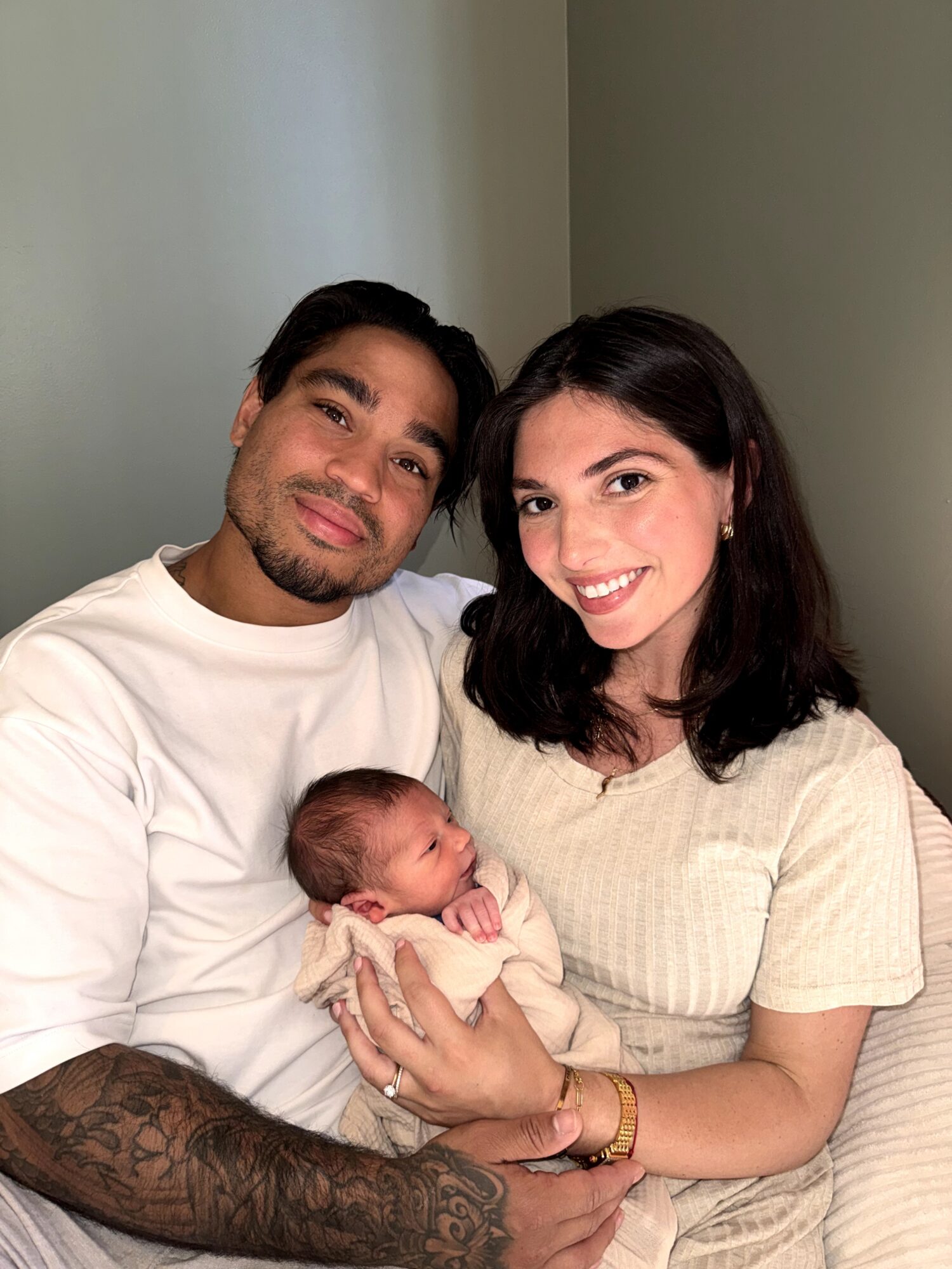 A family of three with a man, woman, and newborn baby, smiling and sitting together indoors.
