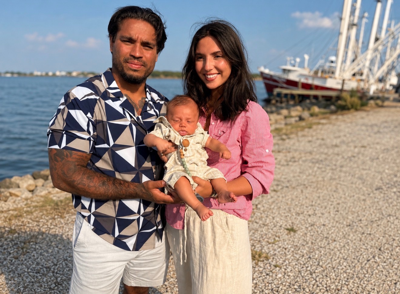 Family of three by the water with boats in the background, smiling, holding a baby, sunny day.