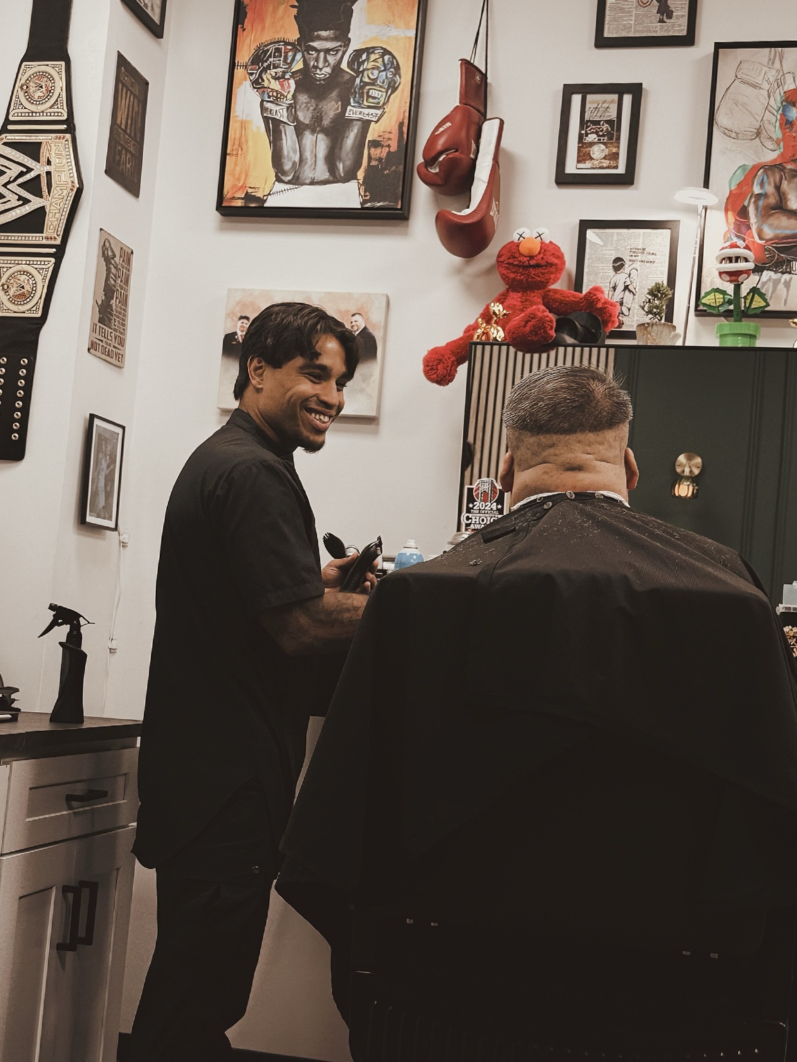 Two men in a room with framed pictures, a red stuffed animal, and a black cabinet, one smiling and the other with back to camera.
