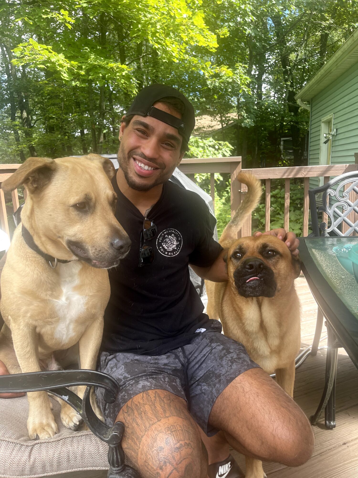 Smiling man with two dogs on a wooden deck surrounded by trees and a house, sunny day.