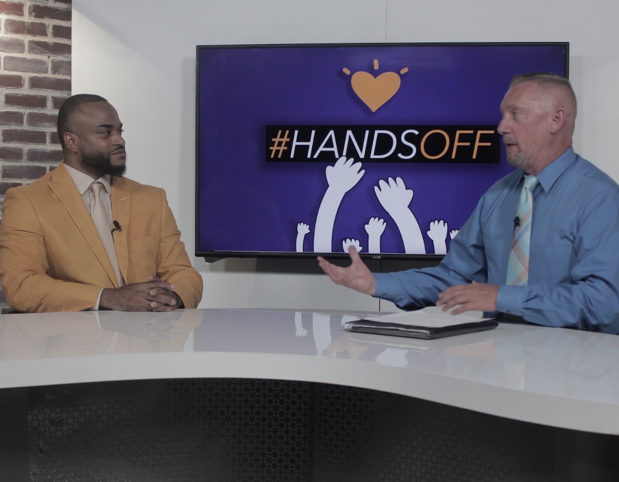 Two men sit at a white table in a studio, discussing with a large screen behind them displaying a hashtag and raised hands.