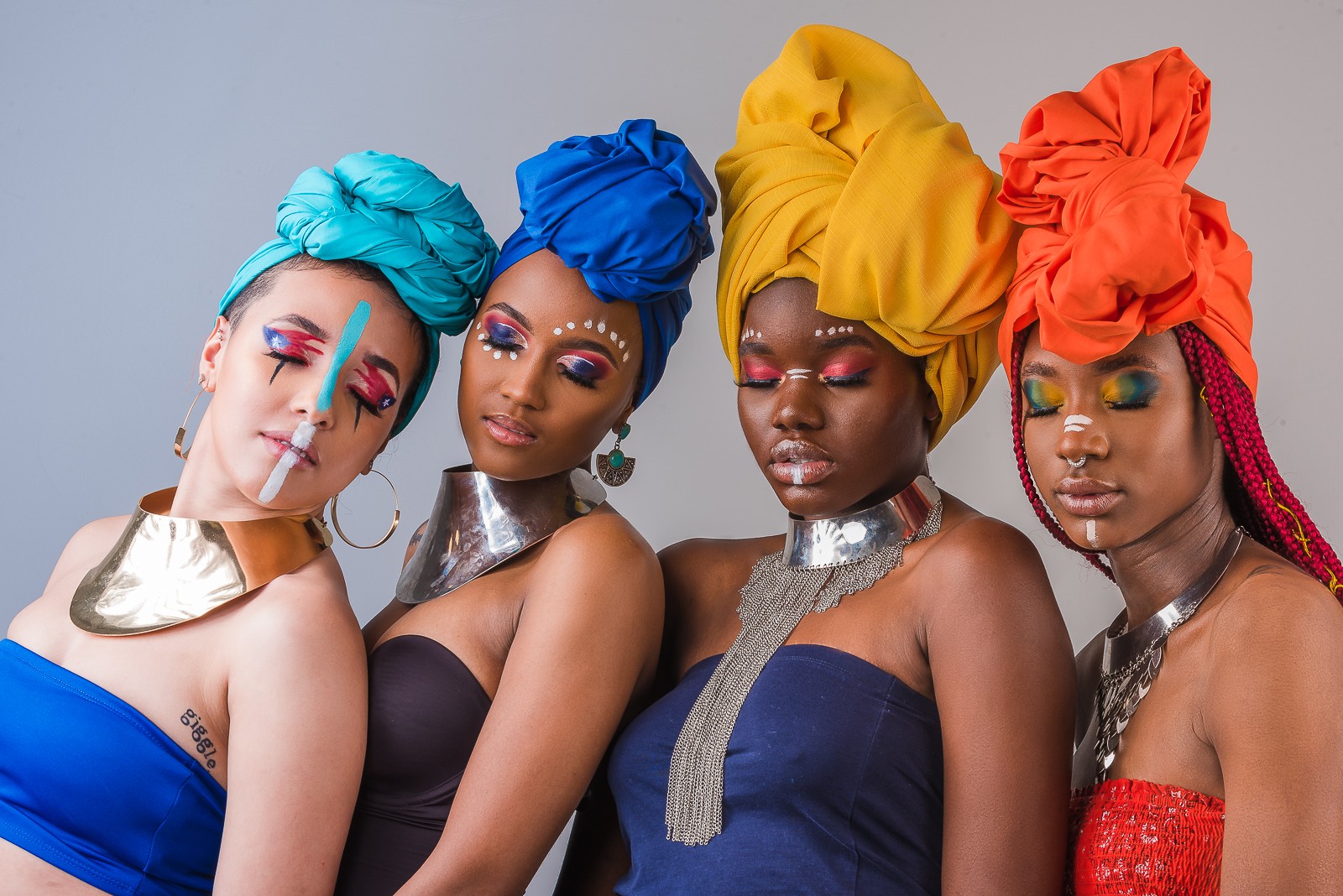 Four women with colorful headwraps and vibrant makeup pose together, wearing strapless dresses and metallic accessories.