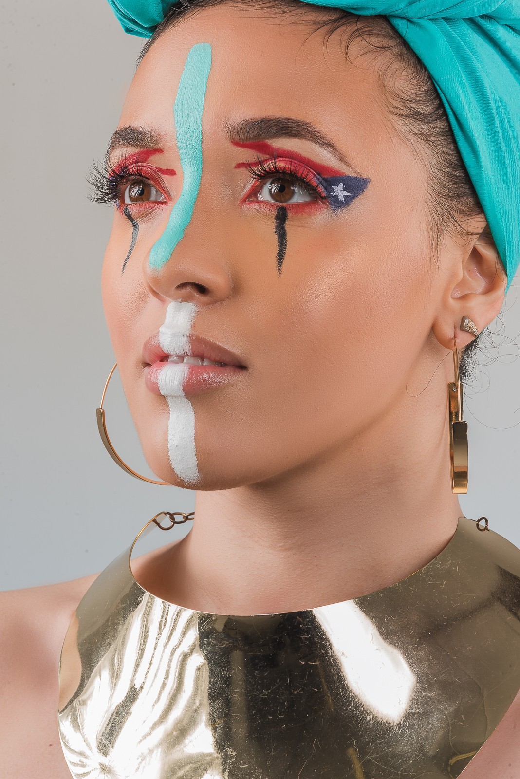 Close-up of a woman with colorful face paint, wearing a teal headscarf and gold earrings, looking to the side.