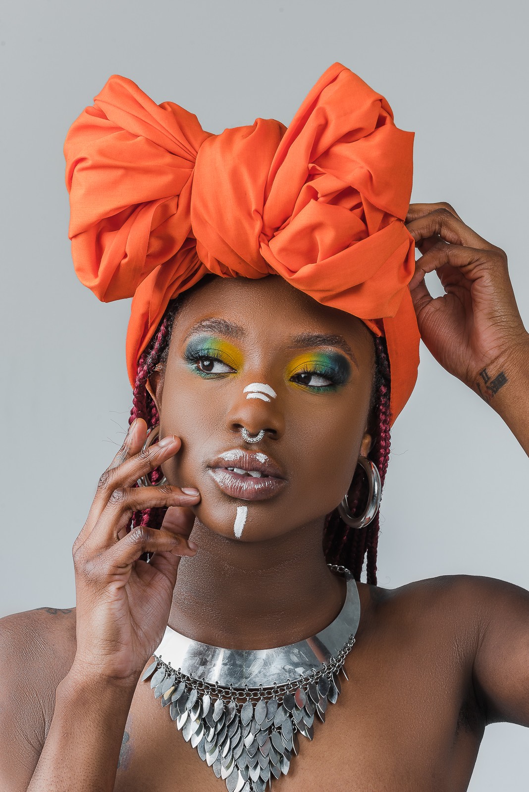 Woman with large orange headwrap, colorful makeup, and silver jewelry, touching her face with one hand.