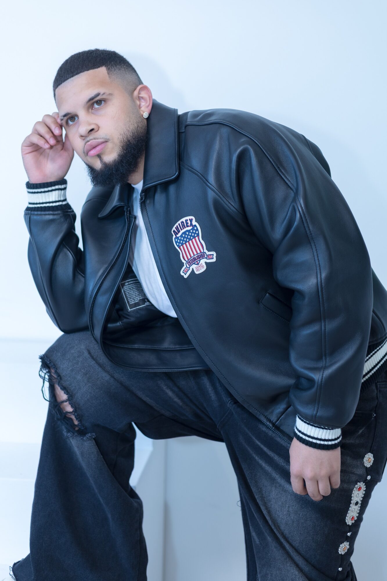 Young man with beard wearing a black jacket with a patch, sitting and leaning forward, looking at the camera.