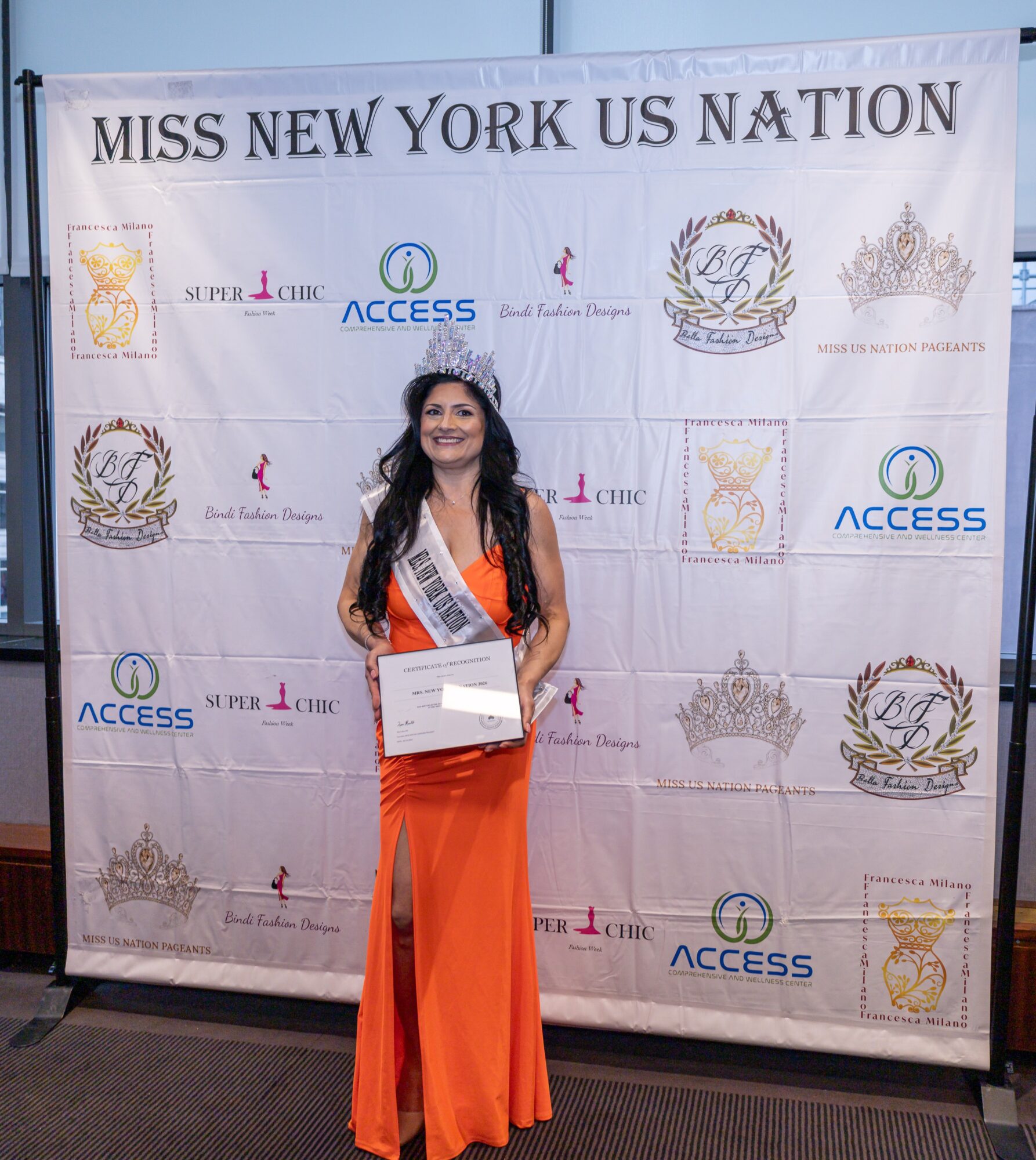 Woman in orange gown holding a certificate, standing in front of a Miss New York banner with logos and text.