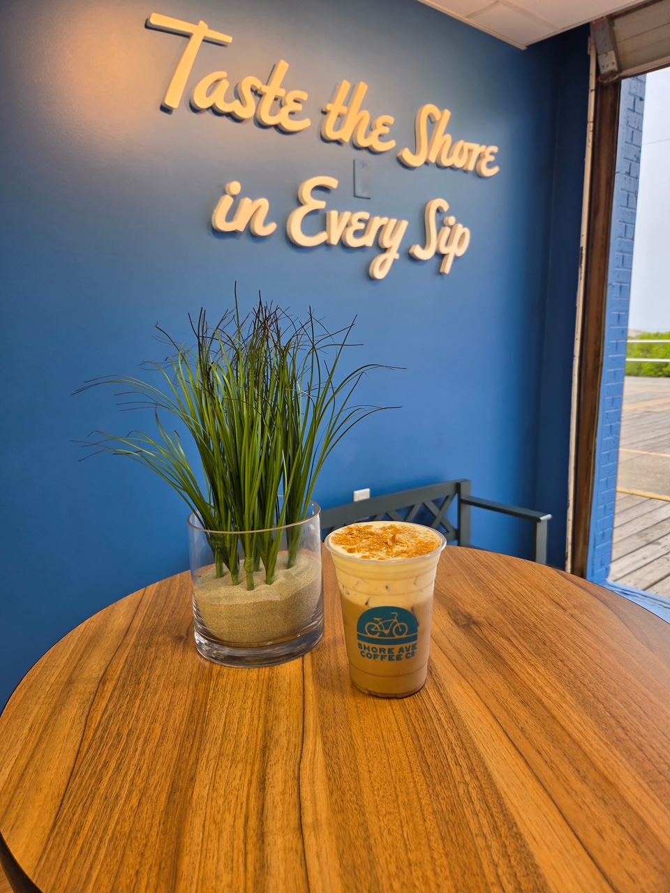 A table with a potted plant and a cup of coffee inside a cafe with a blue wall and a window, text on wall reads 'Taste the Shore in Every Sip'.