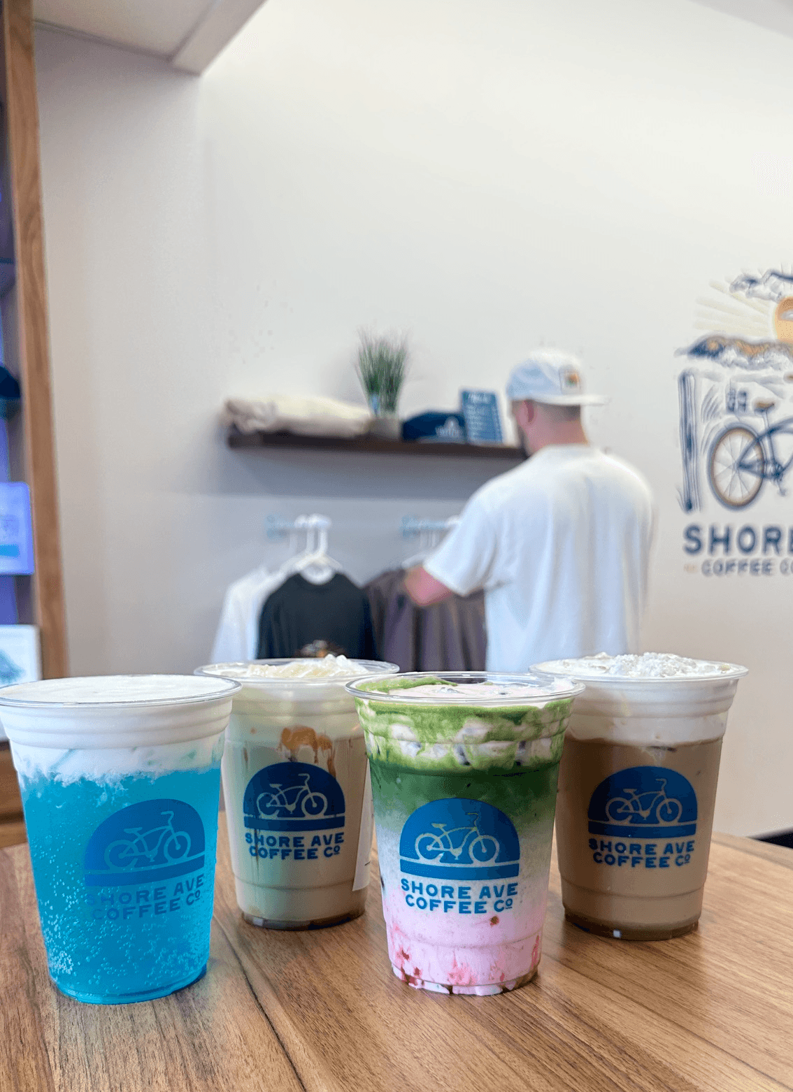 Four cups with beverages on a wooden table, with a person in a white shirt and cap in the background, and a shelf with plants and items. 20 words.
