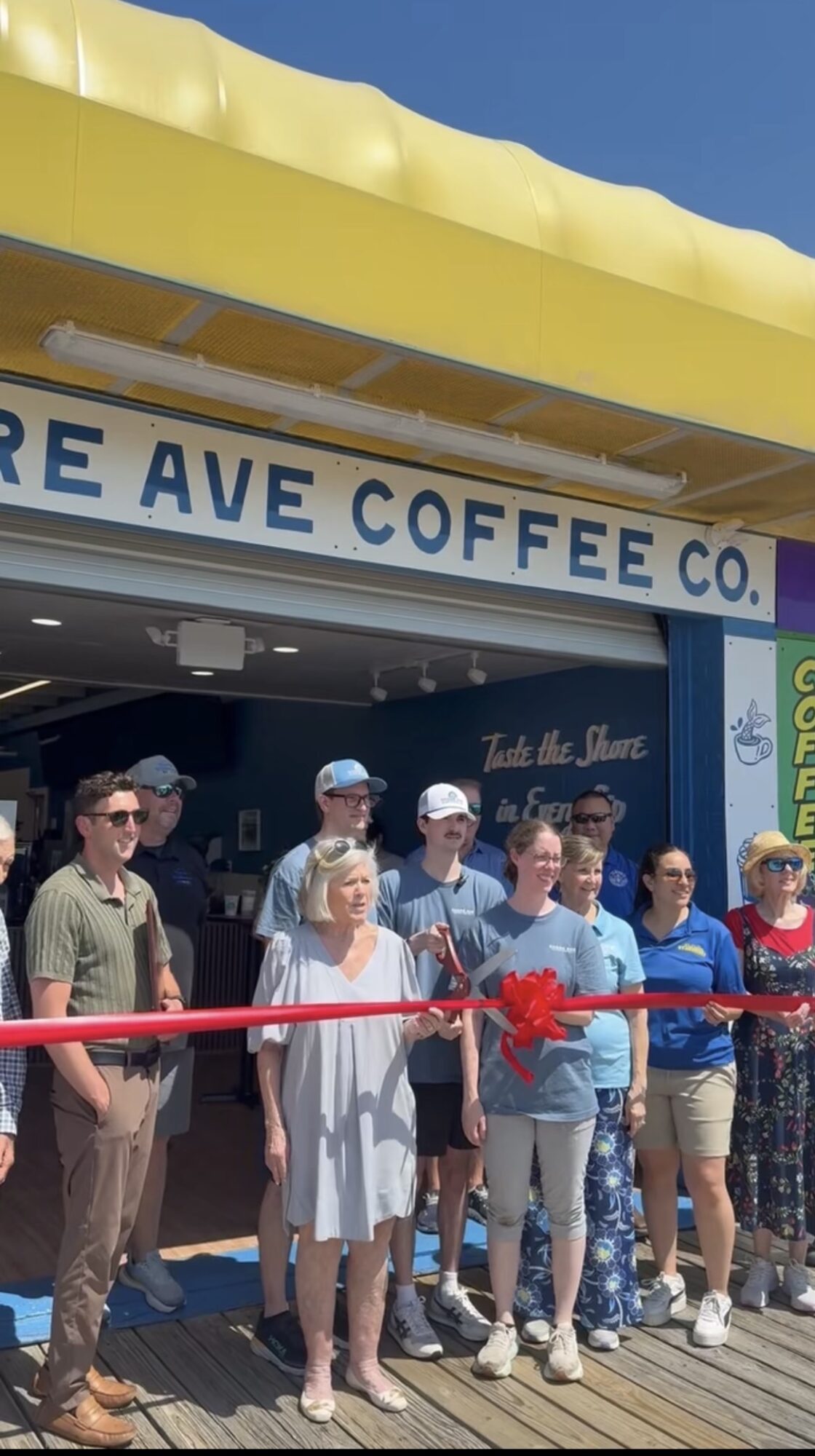 Group of people standing outside a coffee shop with a red ribbon, preparing for a ribbon-cutting event.